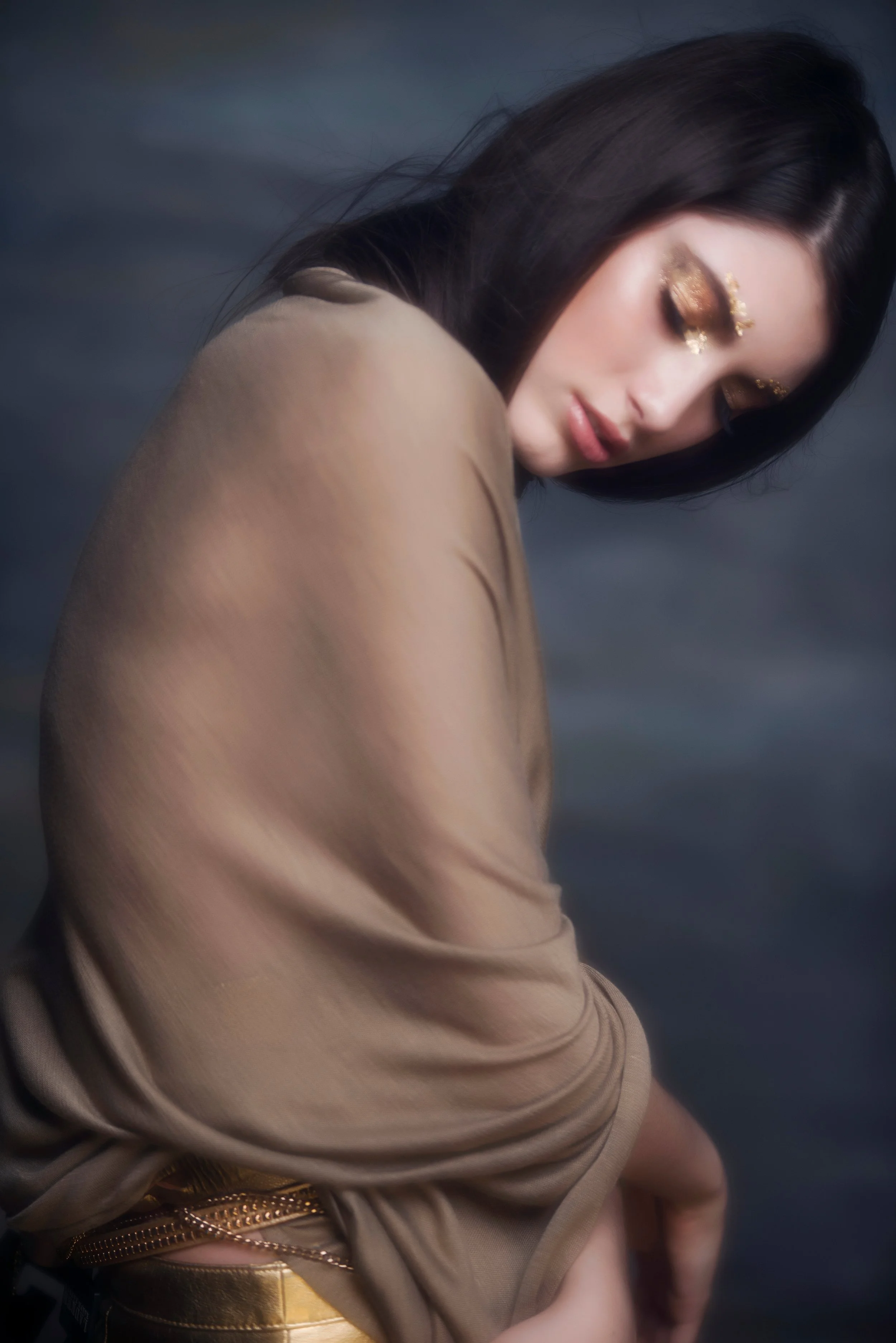A woman with long dark hair and gold makeup, wearing a beige top, poses with her eyes closed against a dark background.