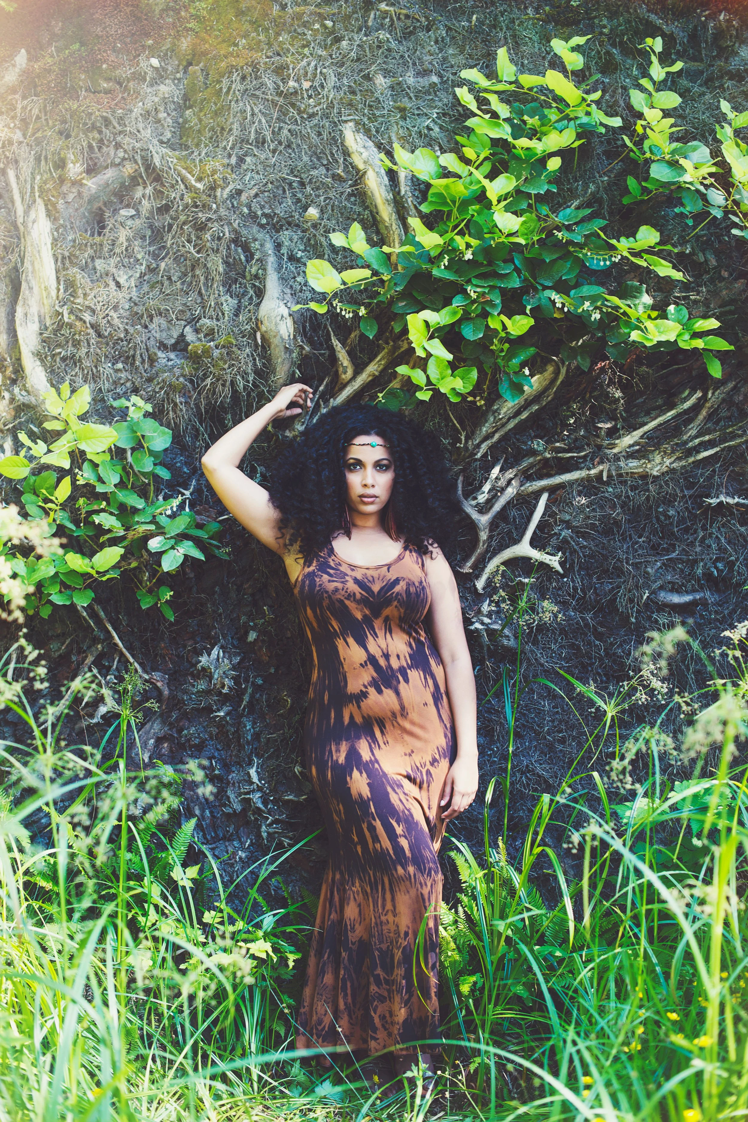 A woman with dark curly hair and wearing a tiger print dress stands with her back against a tree in a lush green forest, surrounded by foliage and tall grass.