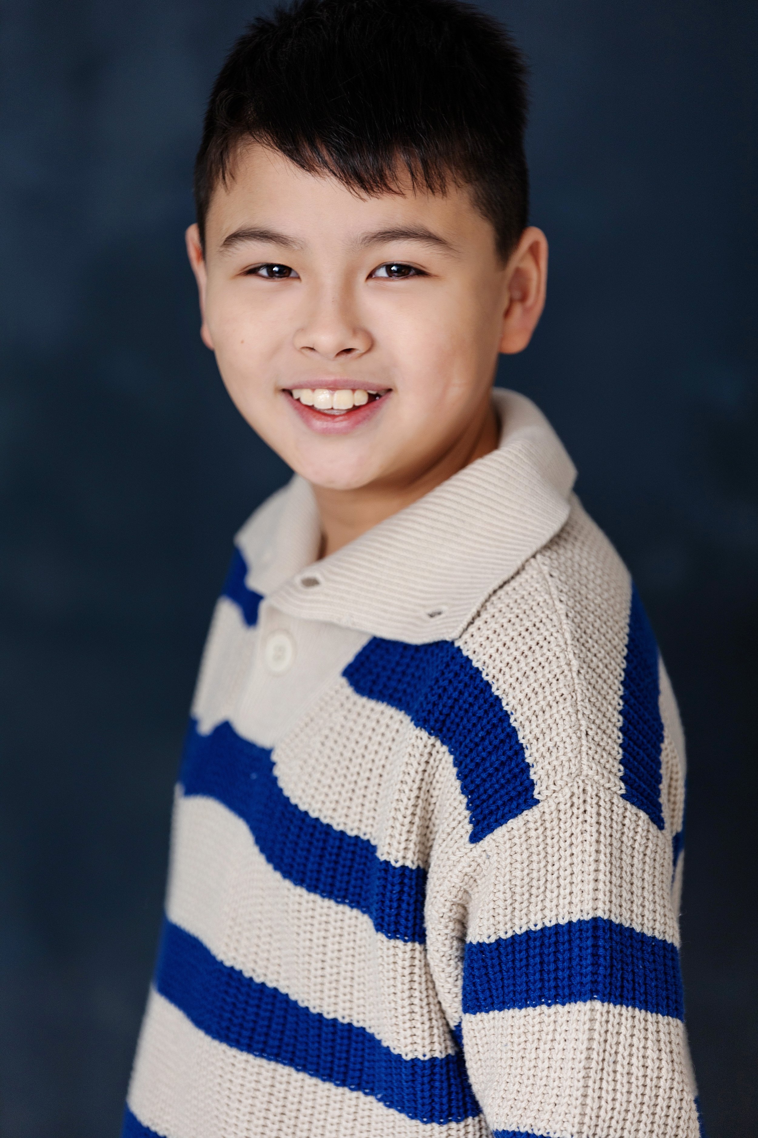 A young boy with dark hair smiling and wearing a beige and blue striped knitted sweater.