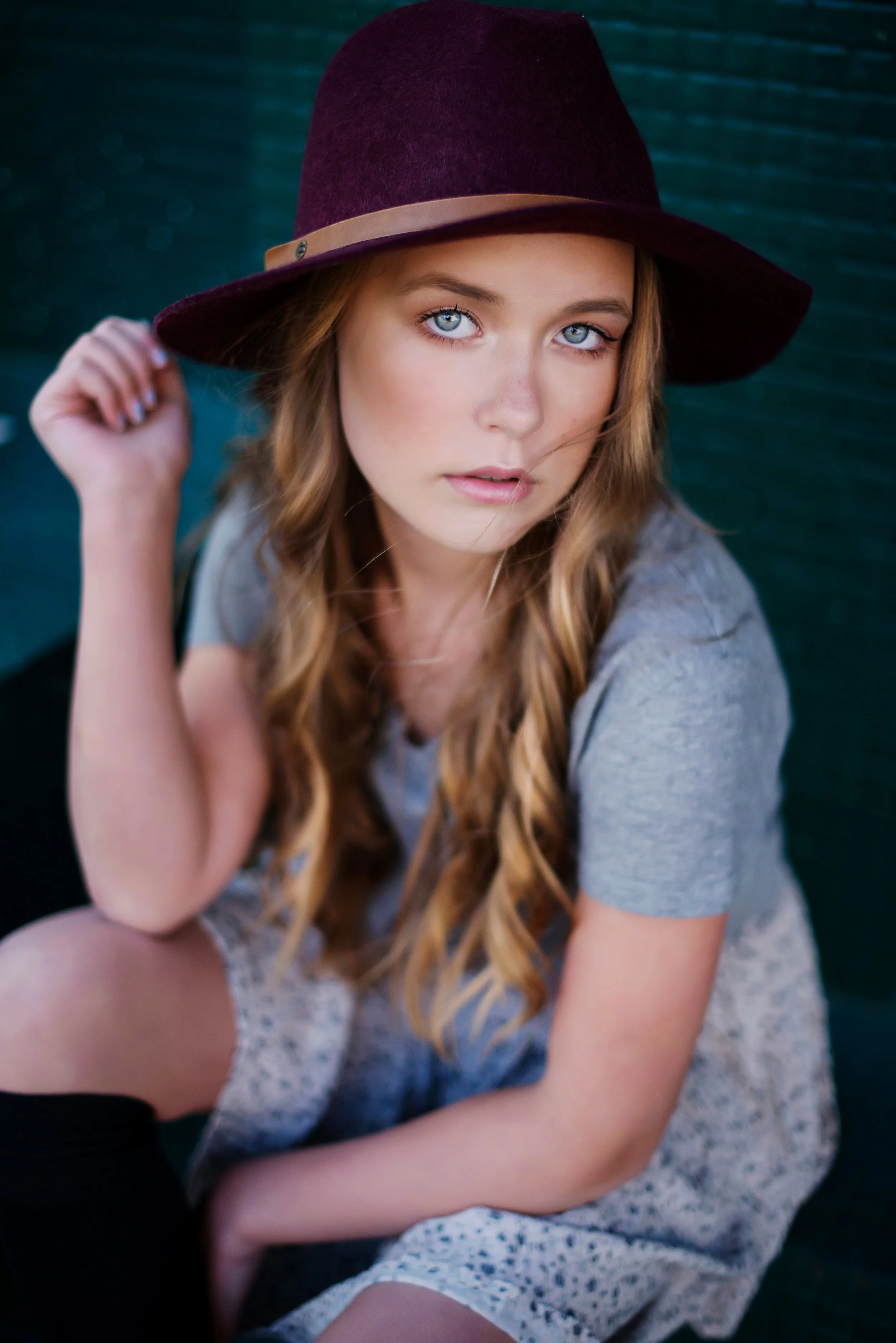 A young woman with long wavy blonde hair and blue eyes wearing a maroon hat with a beige band, looking directly at the camera, sitting against a dark textured background.