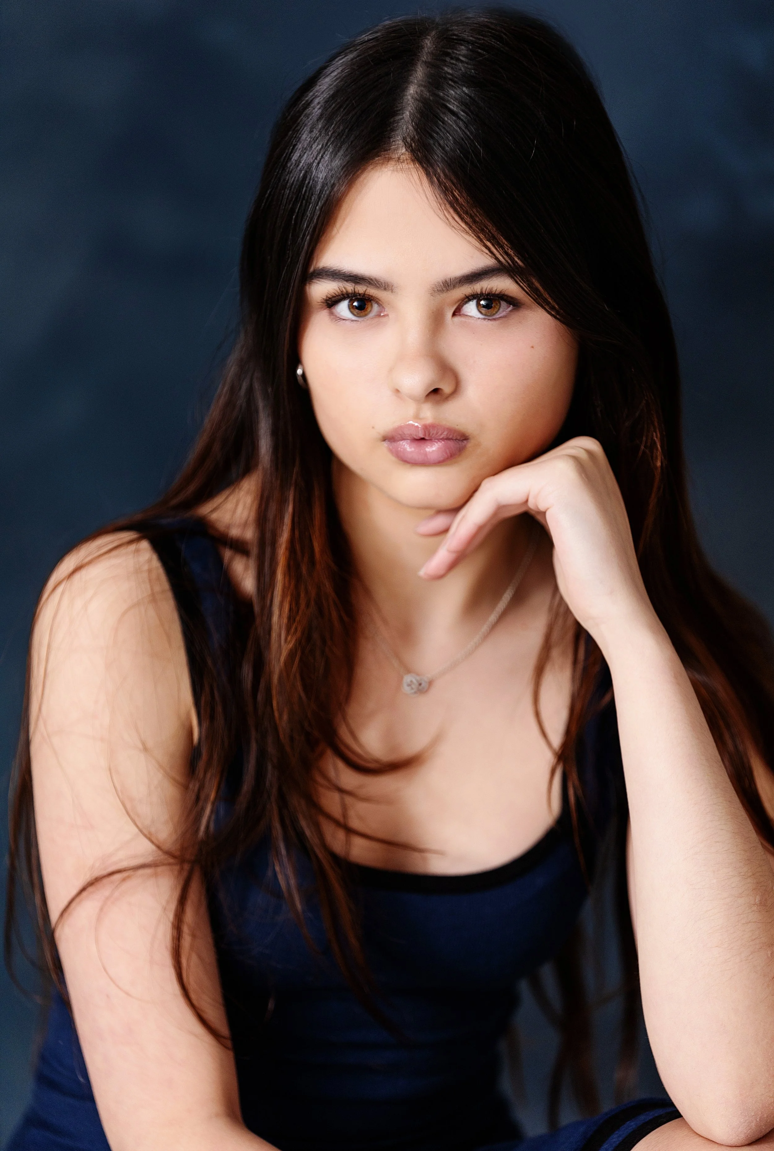 Portrait of a young woman with long dark hair, wearing a black top, resting her chin on her hand, with a dark background.