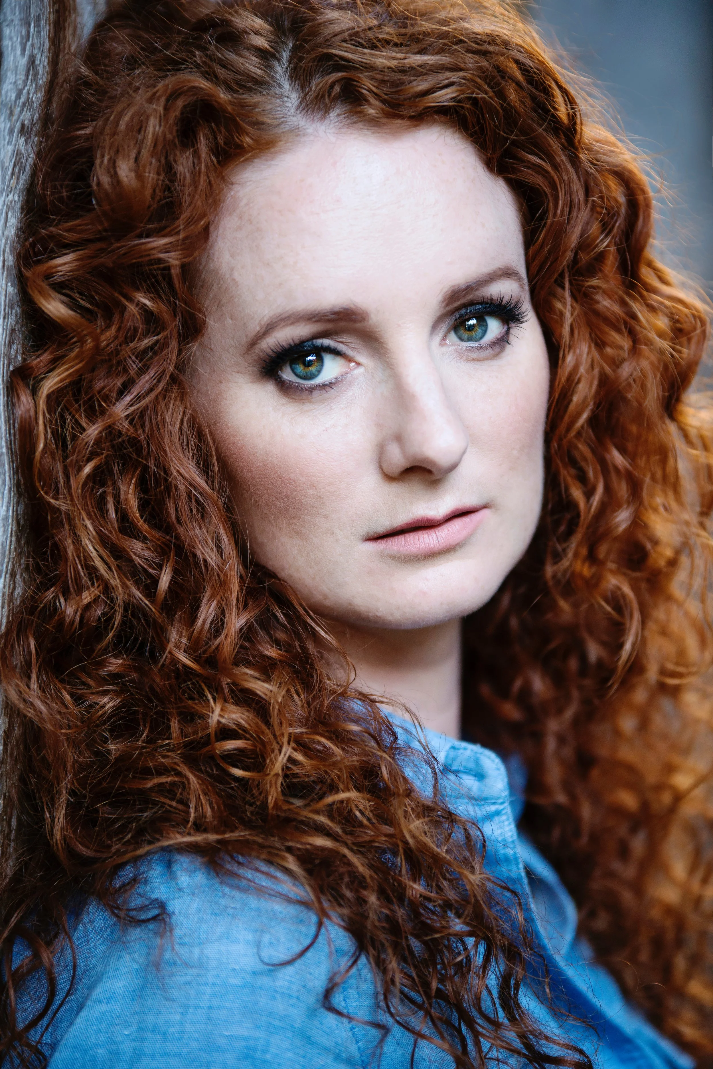A close-up of a woman with curly red hair and blue eyes, wearing a light blue shirt, leaning against a wooden surface.