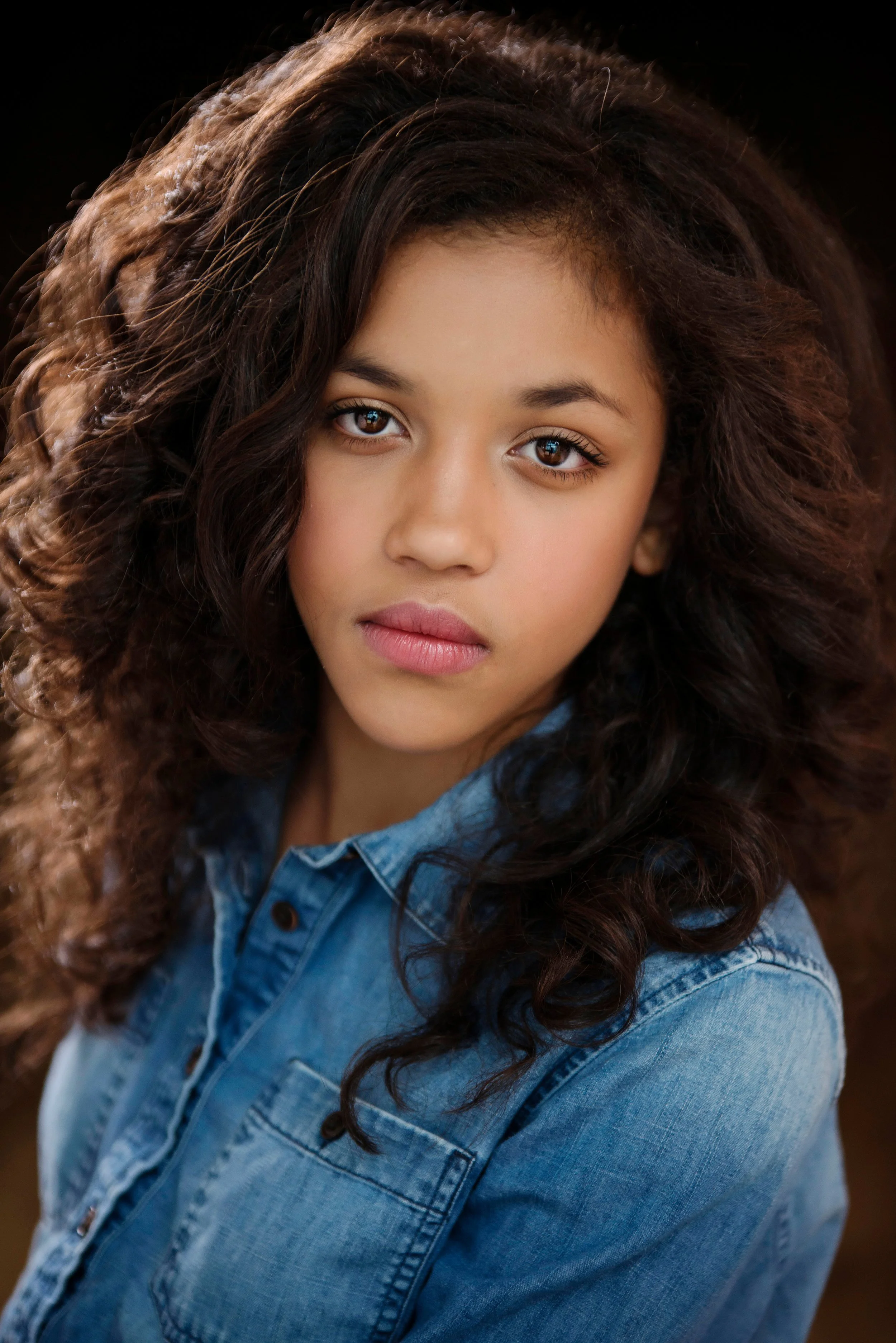 A young girl with curly dark brown hair and brown eyes, wearing a denim jacket, looking into the camera with a neutral expression.