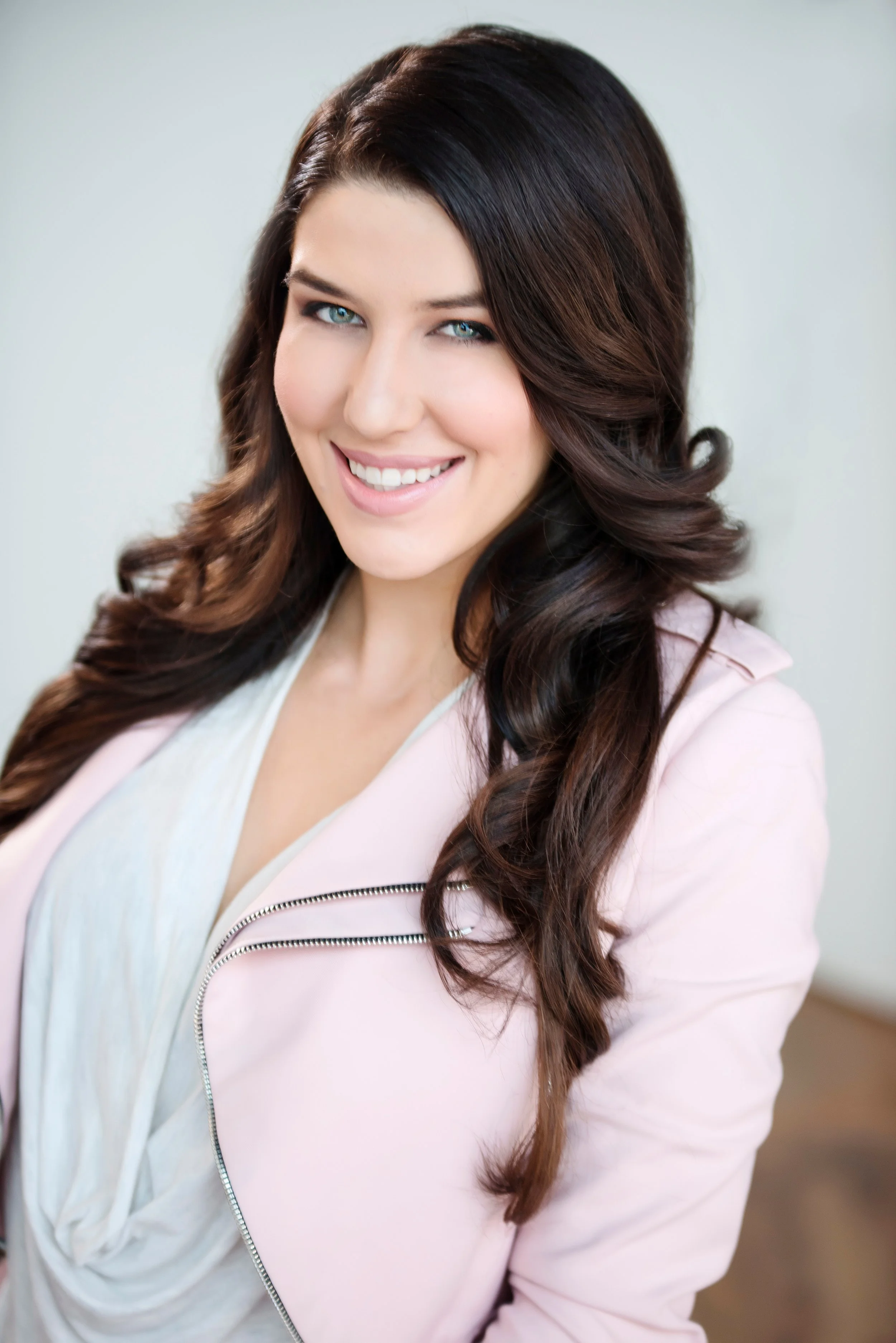 A young woman with long, dark, wavy hair and blue eyes smiling wearing a light pink jacket over a white top.