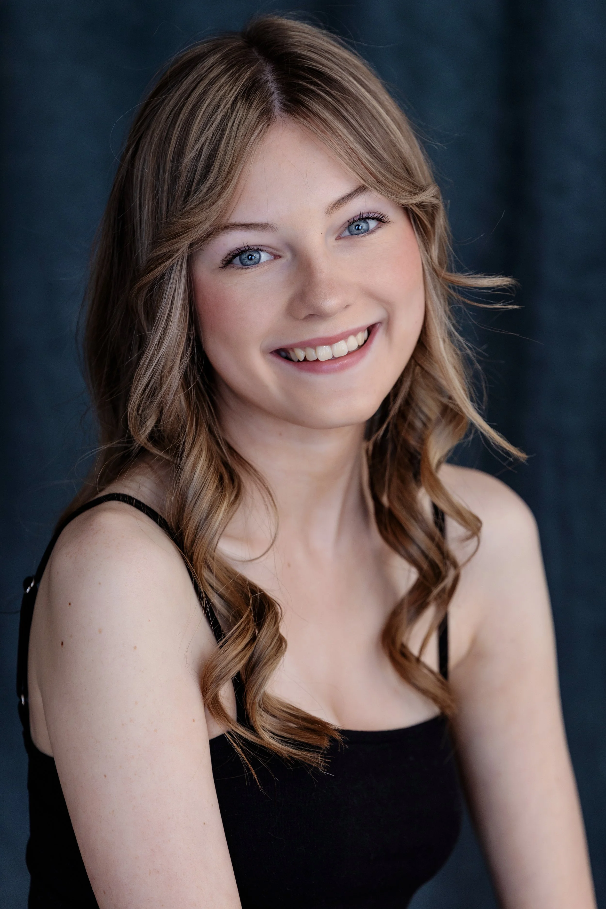 Close-up portrait of a young woman with blue eyes and long wavy brown hair, smiling softly, wearing a black strap top, against a dark background.