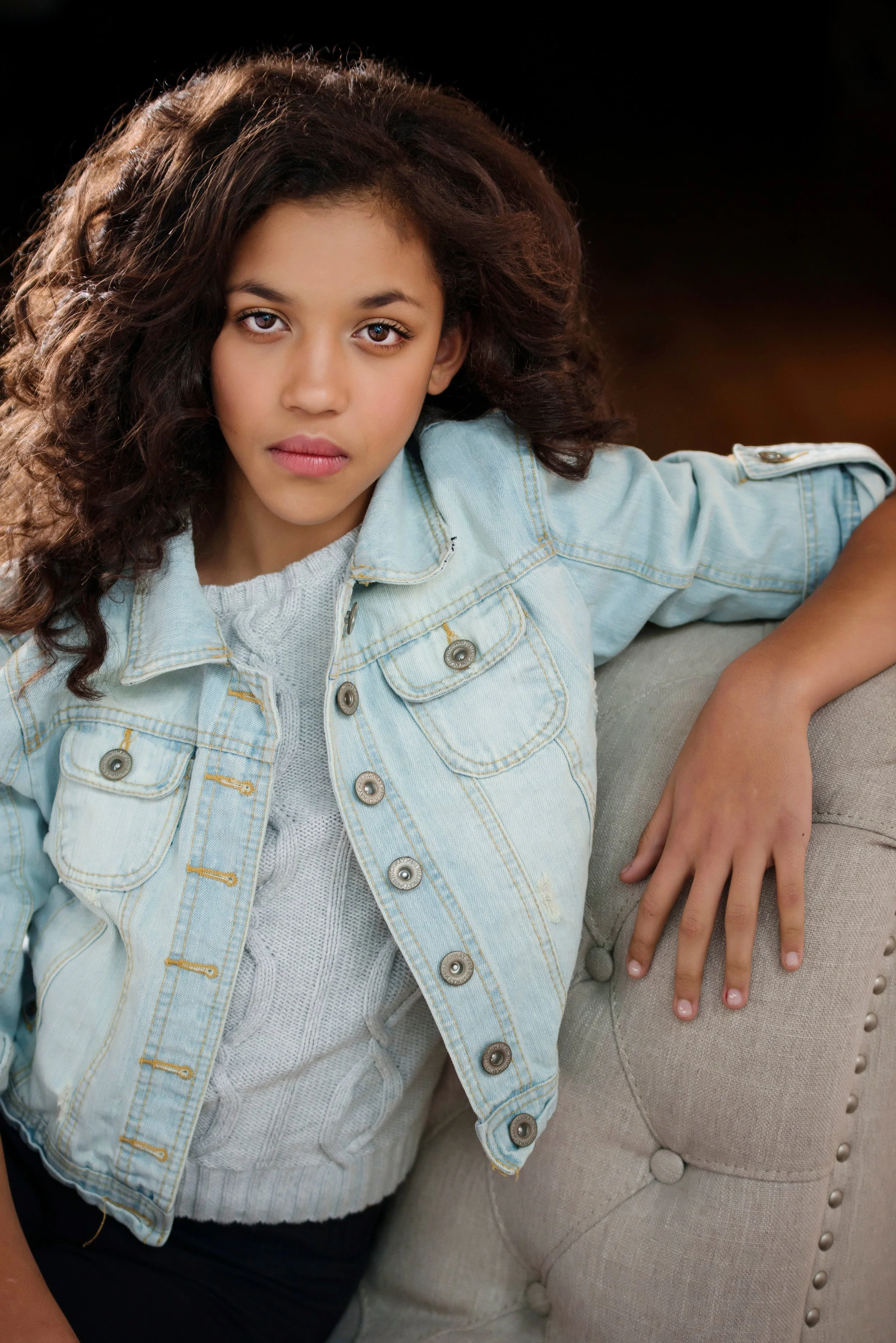 A young girl with curly brown hair and light brown eyes wearing a light blue denim jacket and a white knitted sweater, sitting on a beige upholstered chair with button tufting.