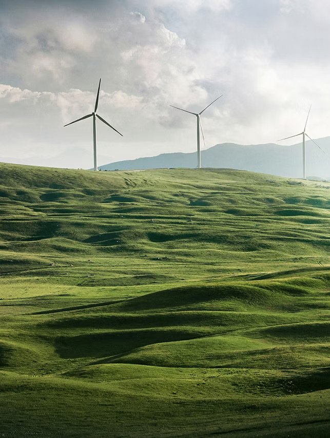 Green rolling hills with three wind turbines and a cloudy sky in the background.
