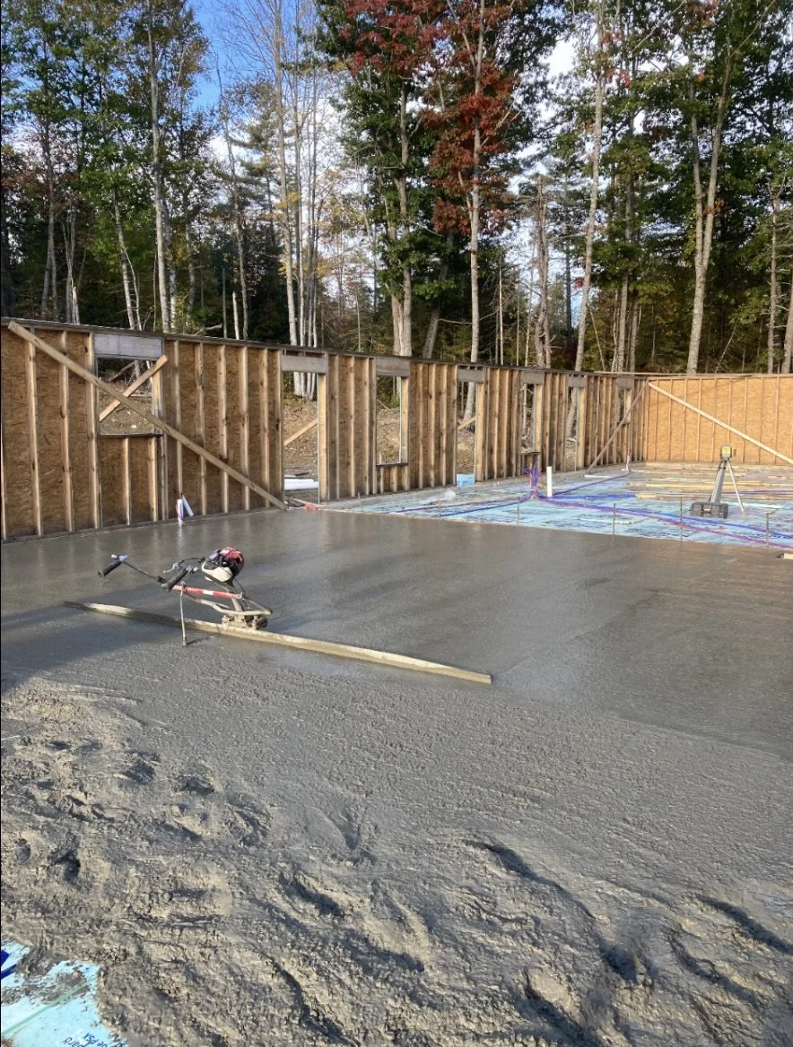 Construction site with freshly poured concrete floor, wooden framing walls, and tools, surrounded by trees.