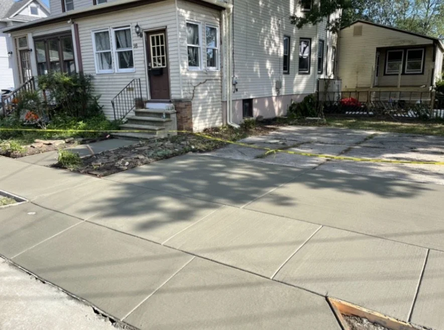 Newly poured concrete sidewalk in front of two houses, with yellow caution tape and shadows of trees.