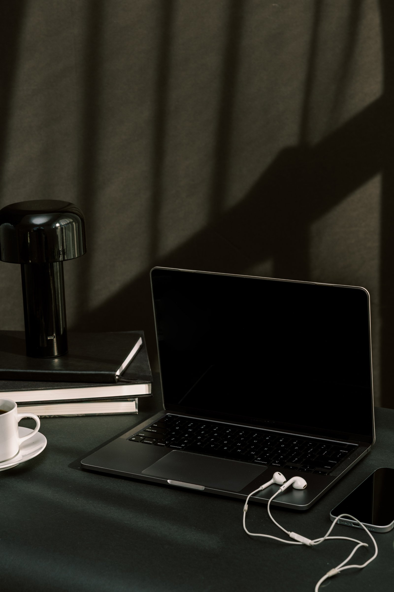 A black laptop on a desk next to a stack of books, a coffee cup, a black table lamp, and a pair of white earphones. The background features a dark curtain with shadows cast by a nearby window.