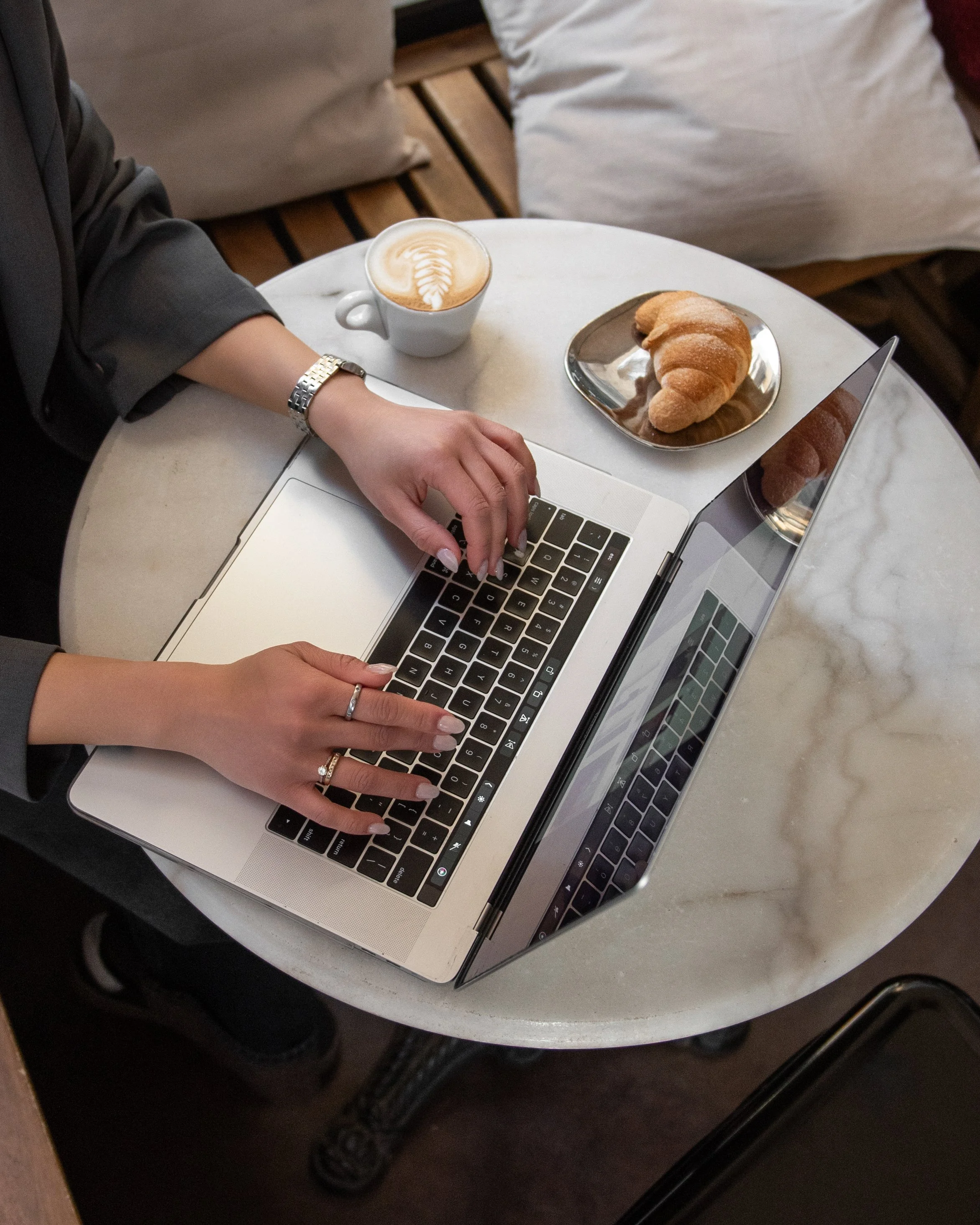A person with a silver watch and rings on their fingers using a laptop with a silver body at a small round table. On the table, there's a white cup of coffee with latte art and a silver tray with a croissant. The table is near a cozy seating area with cushions.