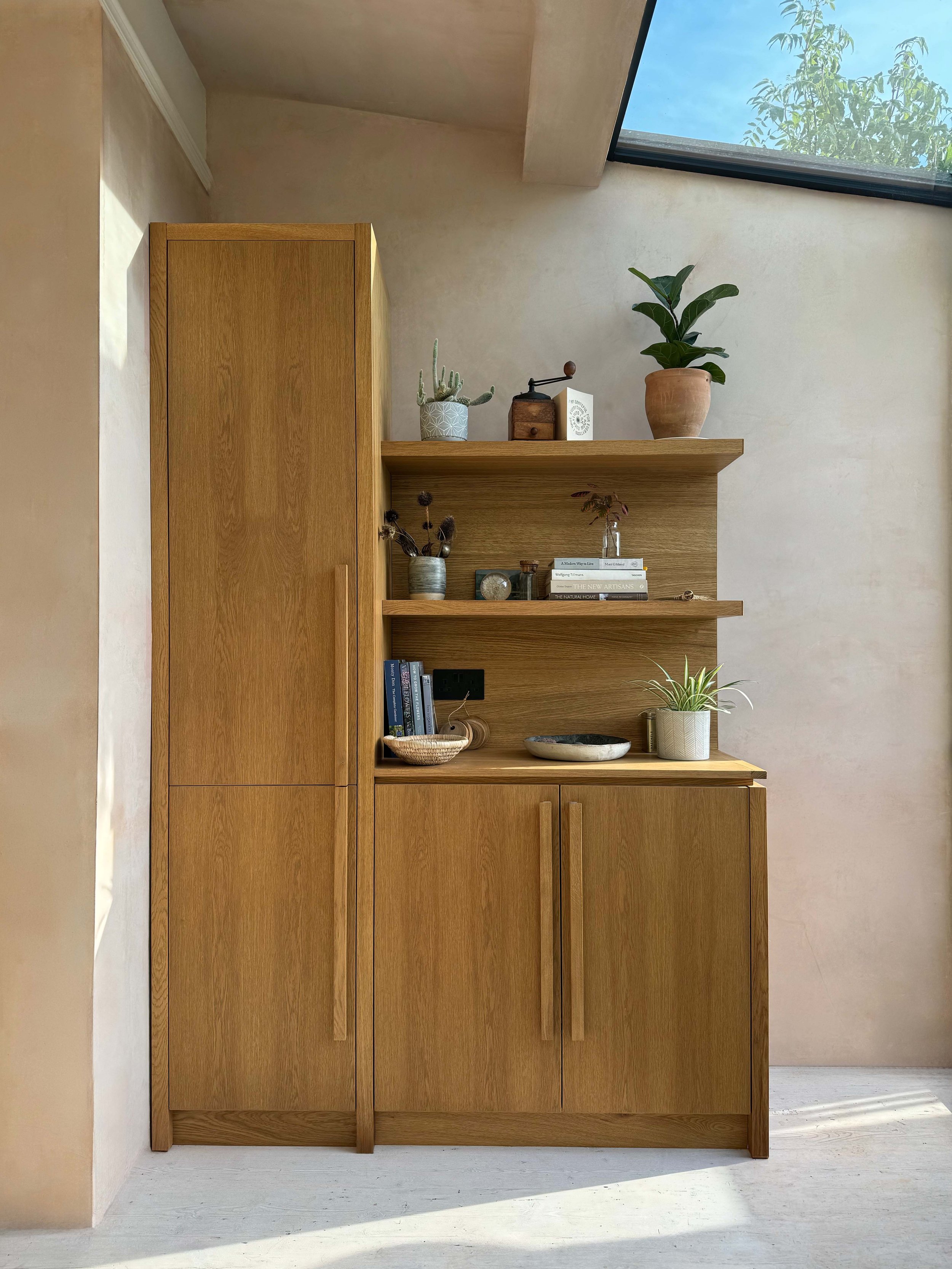 A wooden cabinet with a top shelf displaying potted plants and decorative objects, set against a plain wall with a skylight overhead.