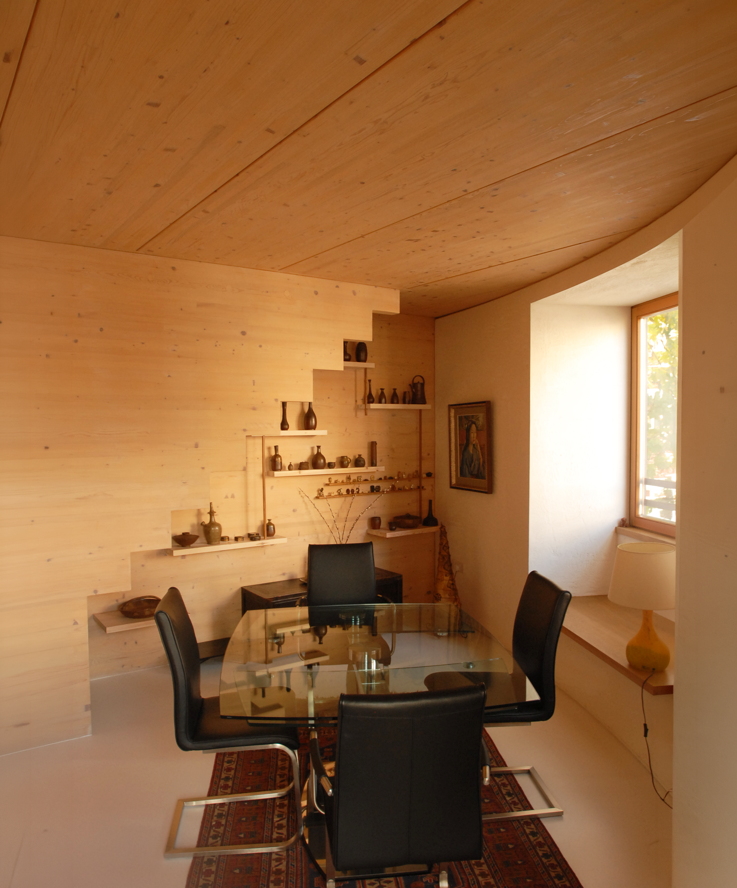 A cozy dinning room with a glass top table surrounded by four black chairs. The room features a wooden wall and ceiling, with two floating shelves displaying vases. There's a window on the right letting in natural light, and a patterned red rug underneath the table.