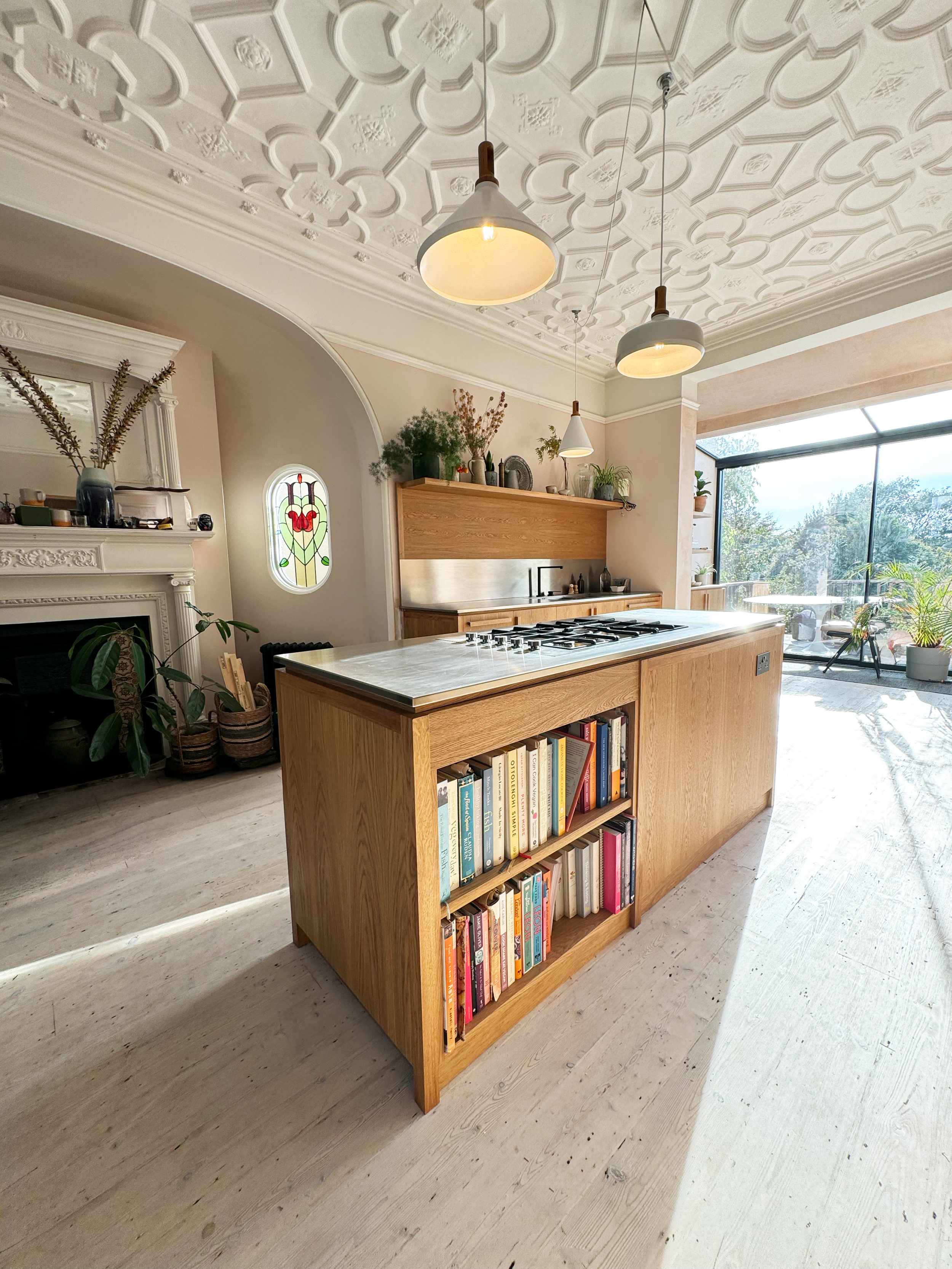 A modern kitchen with a wooden island containing a built-in bookshelf filled with books. The room features a decorative ceiling with white embossed patterns, pendant lights hanging from the ceiling, and a large window letting in natural light. Various plants and decorative items are placed on a wooden shelf above the stove, and there is a stained glass window visible on the wall.