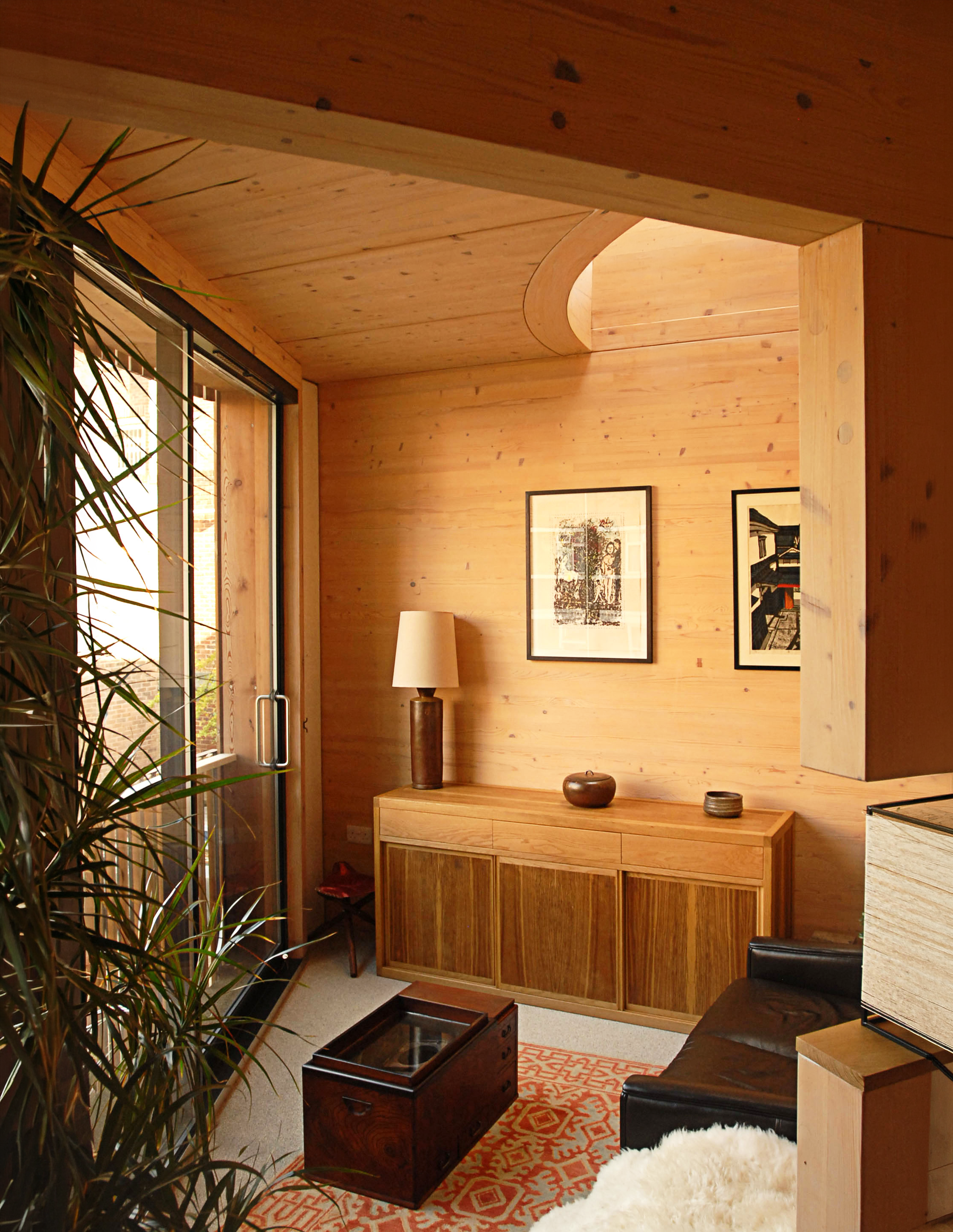 A cozy living room with wooden walls and ceiling. There is a window with a view outside and framed artwork on the wall. The room features a wooden cabinet, a floor lamp, a black sofa, a decorative rug, and a barrel-shaped wooden chest.