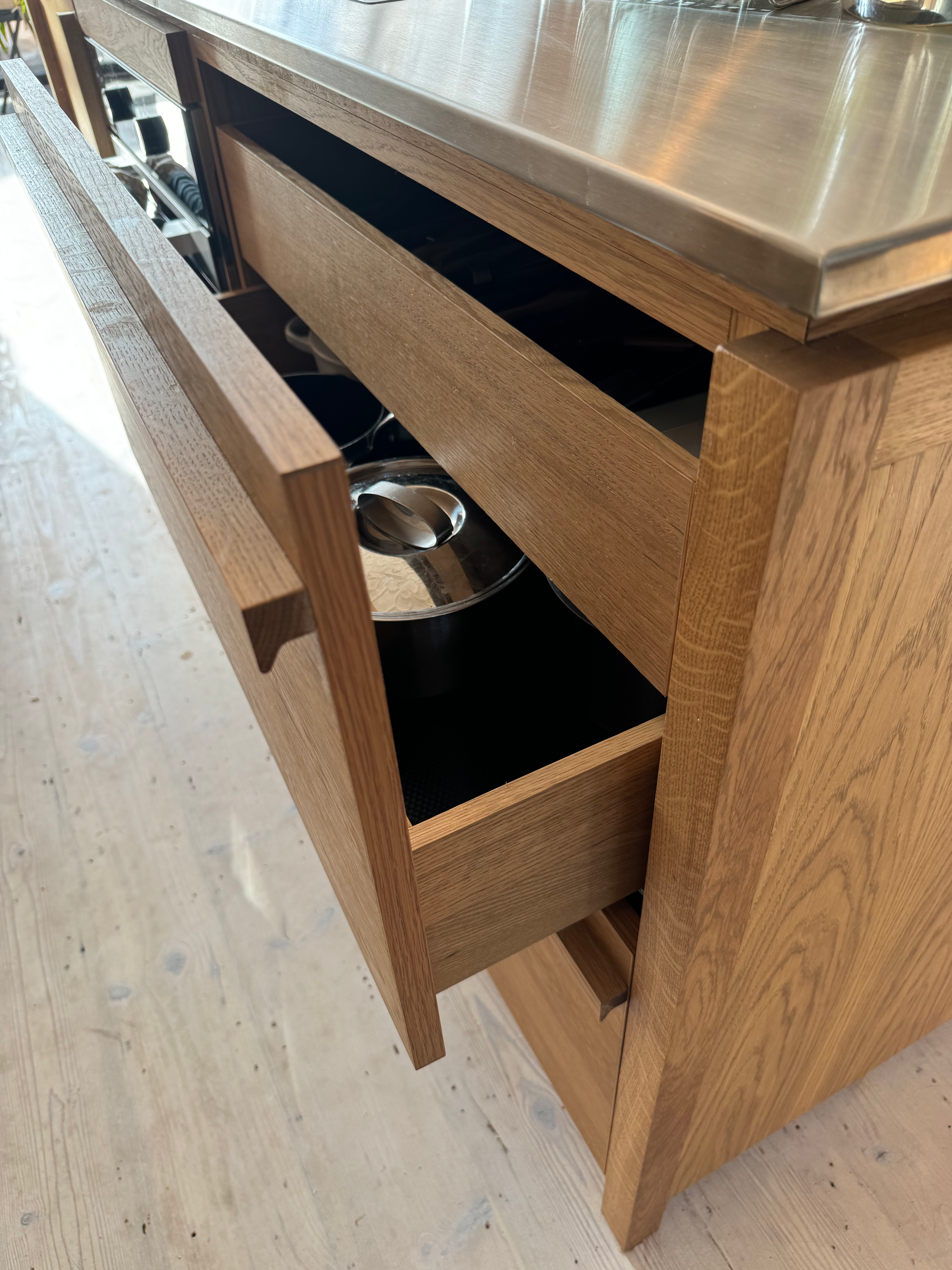 Bespoke Kitchens & Furniture. Open wooden cabinet with stainless steel countertop, showing inside drawers and kitchen utensils.