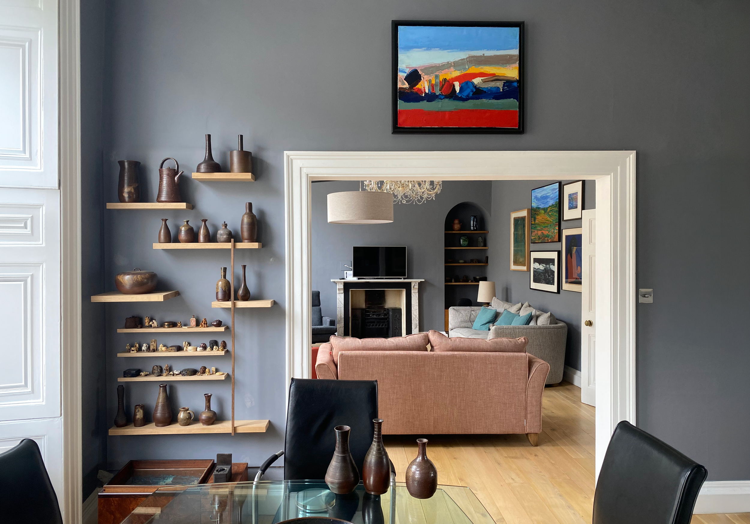 Interior view of a living room with a blue wall, decorative vases on floating shelves, a chandelier, a portrait painting, a fireplace, and sofas with cushions, visible through a doorway.