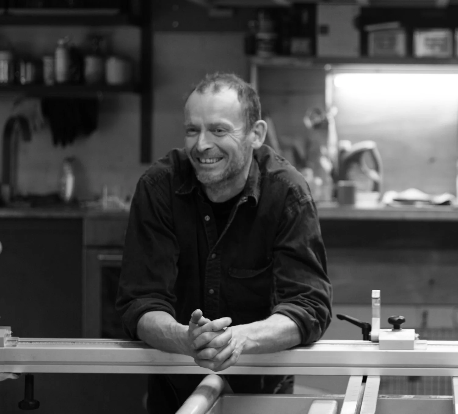 A man smiling with arms resting on a table in a workshop or studio with shelves and tools in the background.
