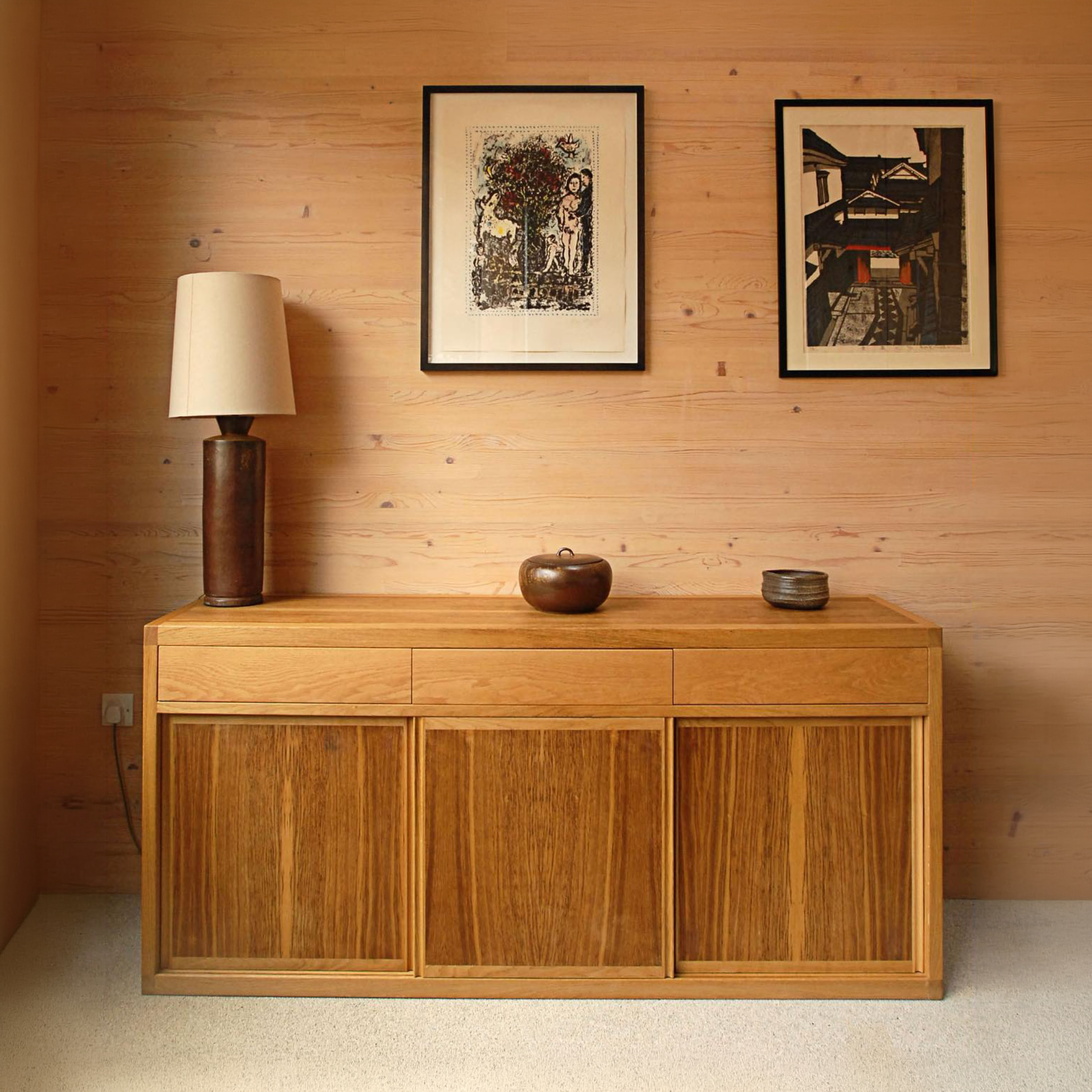 A wooden cabinet with a foldable door, on which sits a collection of pottery, with a wooden wall behind it.