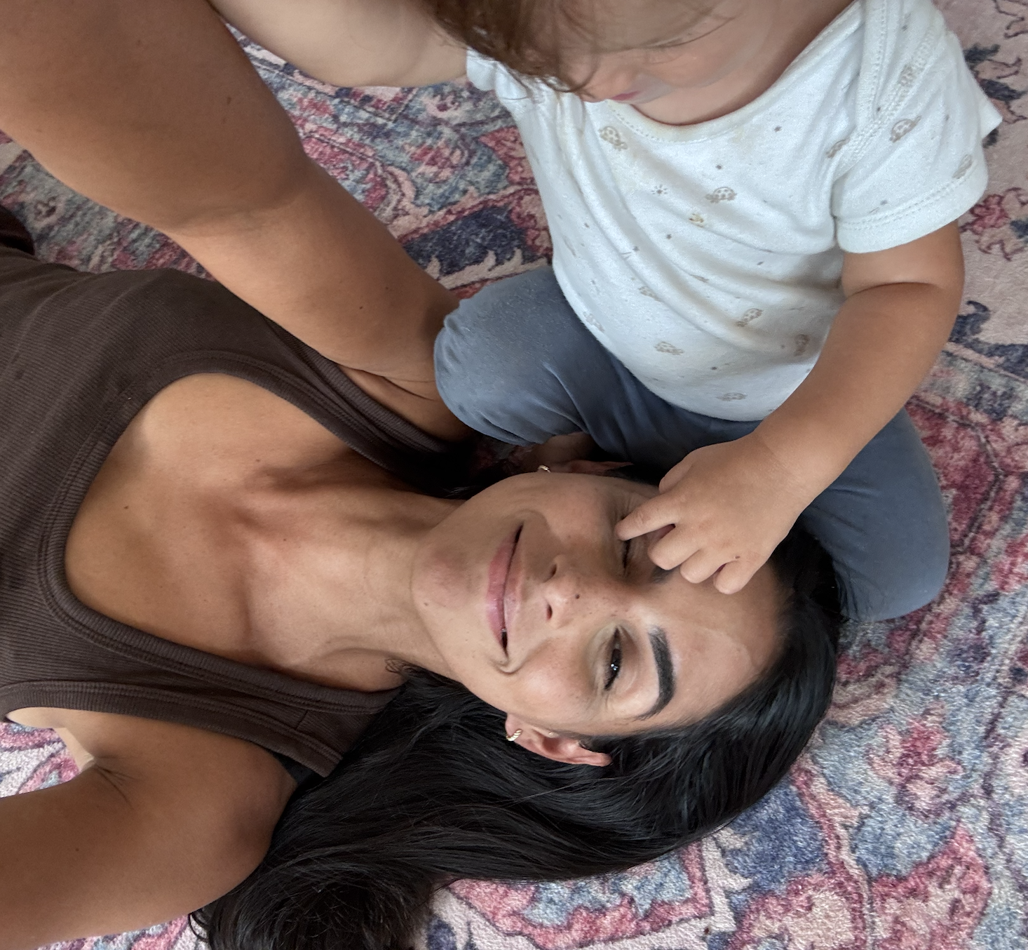 A woman with dark hair and light skin lying on a patterned rug with a young child sitting on her stomach, touching her nose.