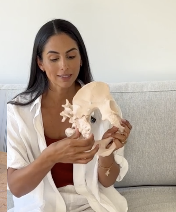 A woman with long dark hair sitting on a light gray couch, holding and examining a human pelvic bone model.