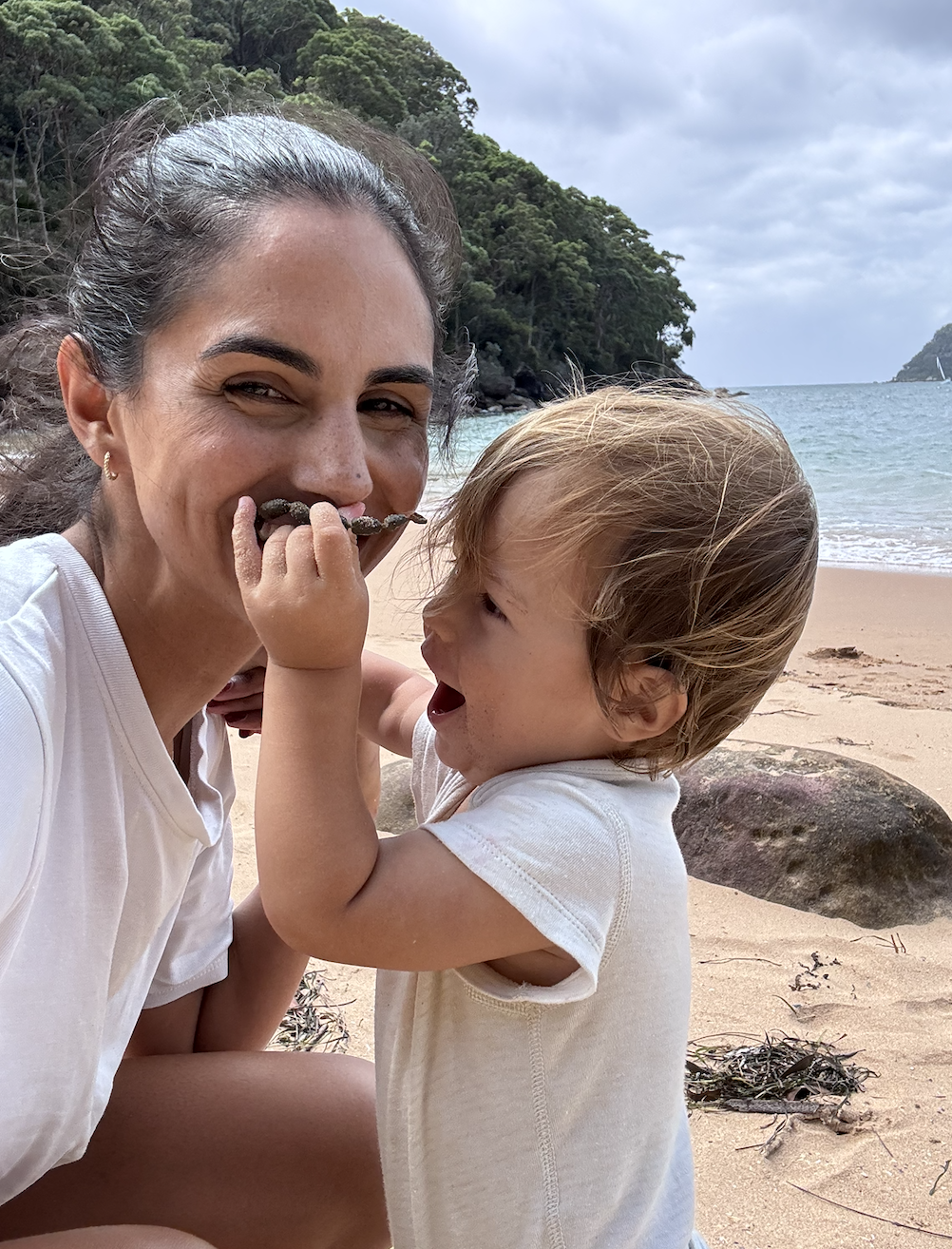 A woman and a young child playing on a beach, with the woman holding a small object in her mouth and the child touching her face, with the ocean, rocky shore, and green cliffs in the background.