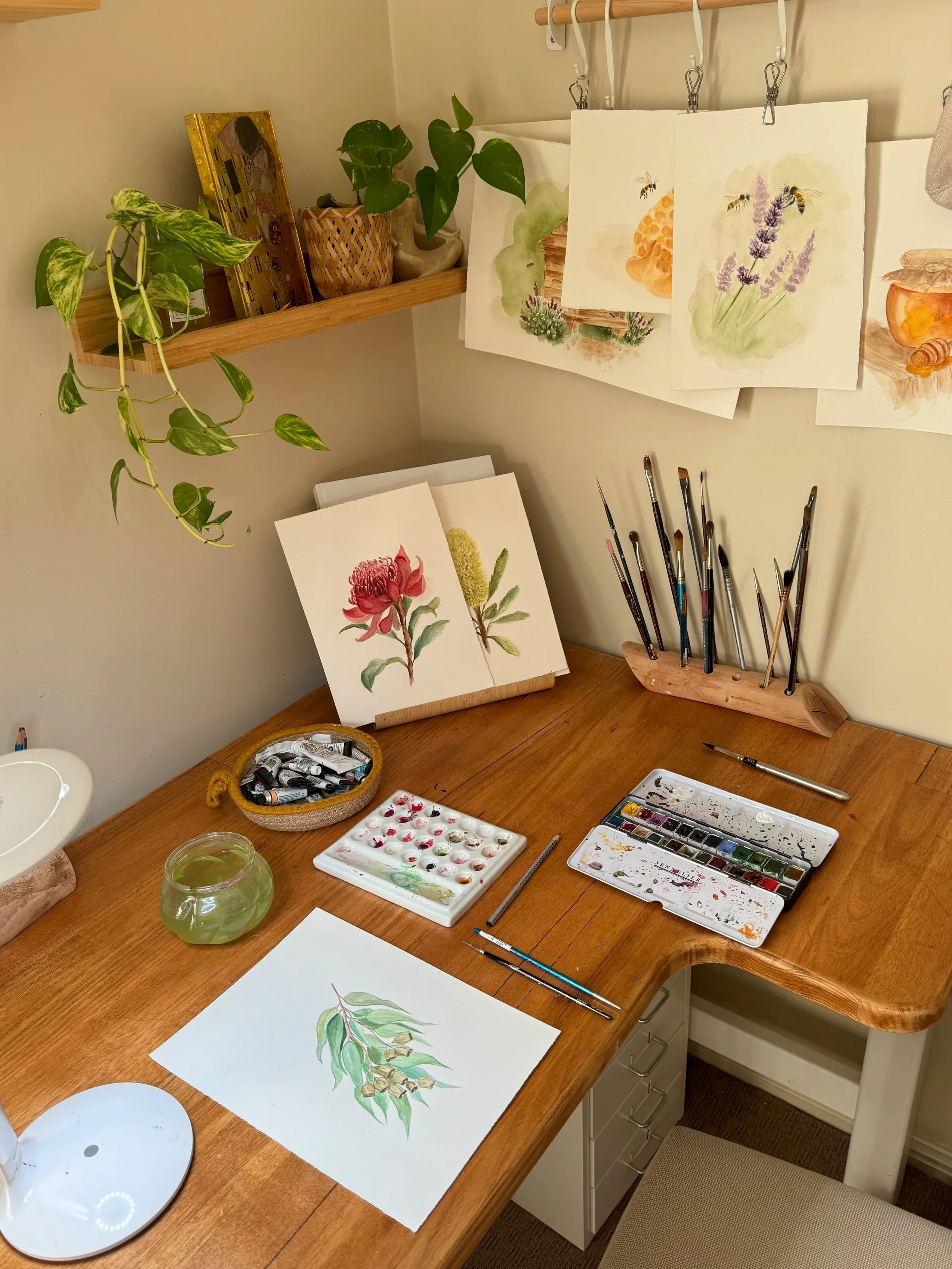 Art supplies and watercolor paintings on a wooden desk and wall shelf, with a set of watercolor paints, brushes, and a small bowl of green liquid.