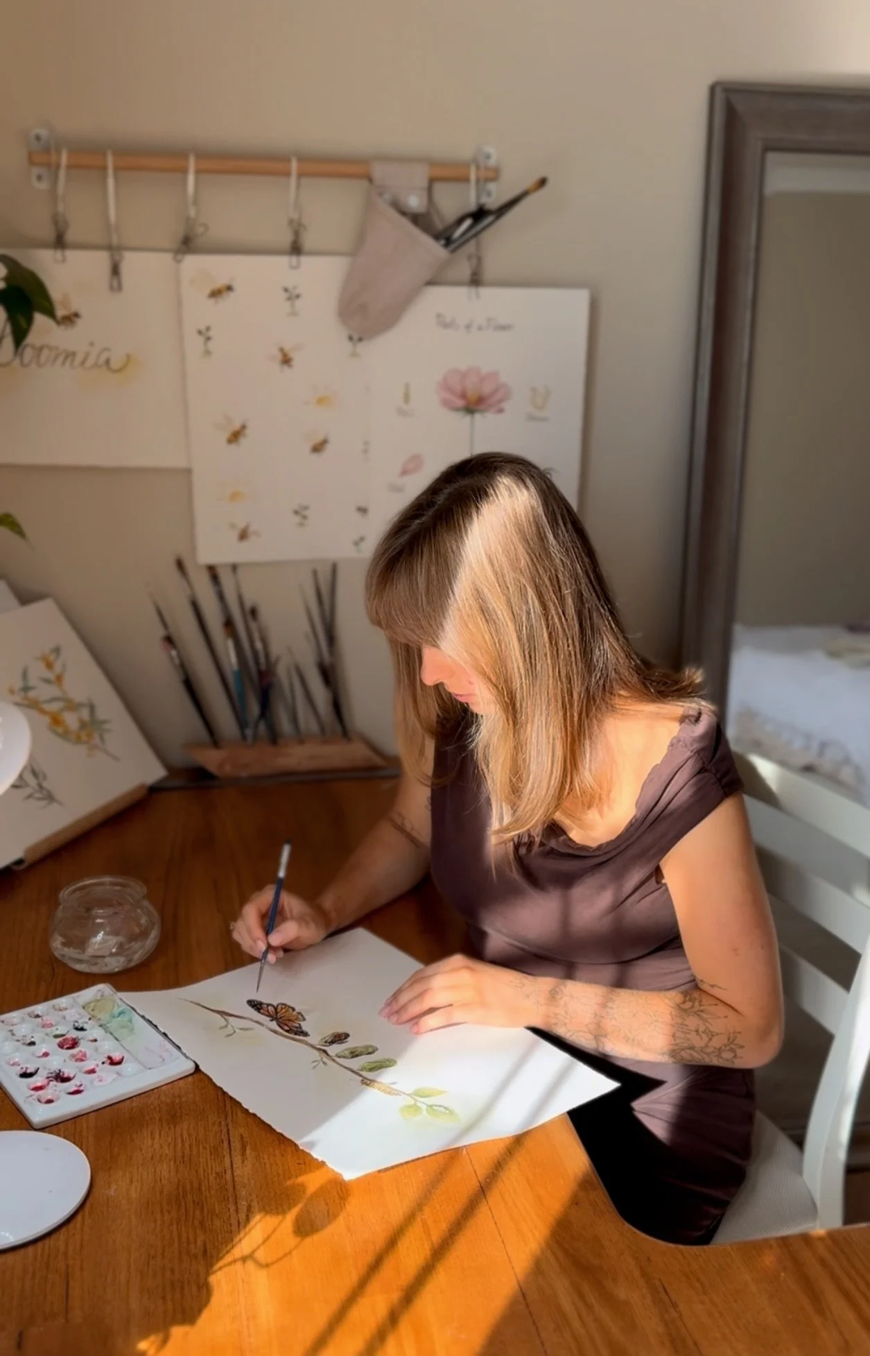 A woman with shoulder-length hair painting a butterfly on paper at a wooden table. Art supplies and watercolor paints are visible nearby. Behind her, art prints and a shadow of a branch are on the wall.