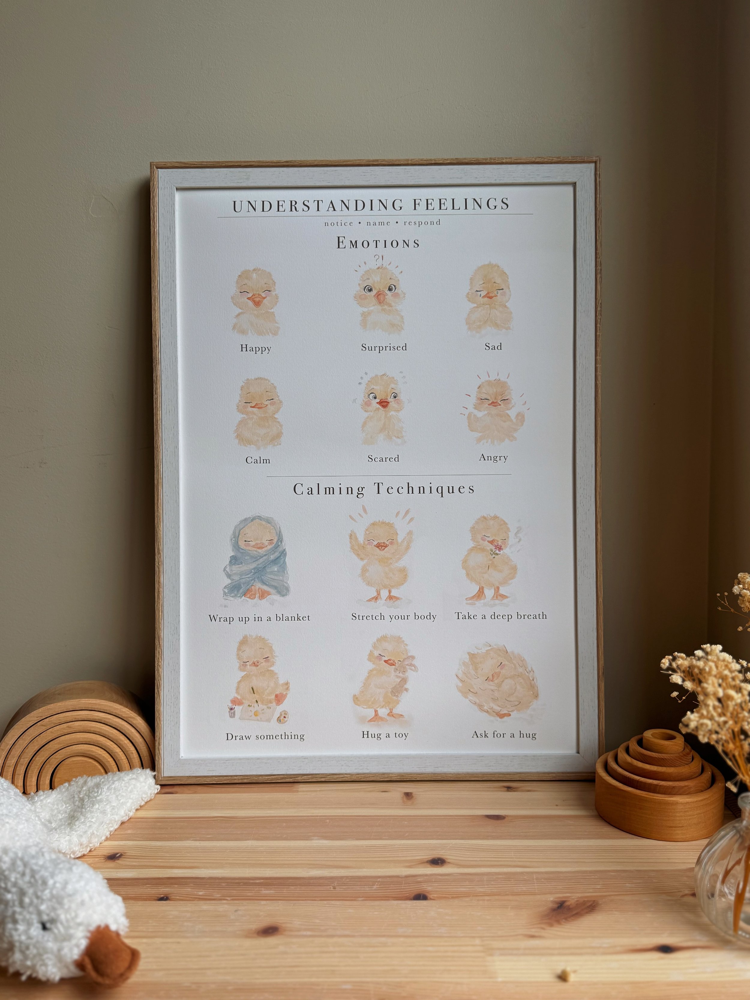 A framed poster titled "Understanding Feelings" with watercolor illustrations of ducklings depicting different emotions and calming techniques, placed on a wooden table with decorative wooden arches and a plush white sheep toy.