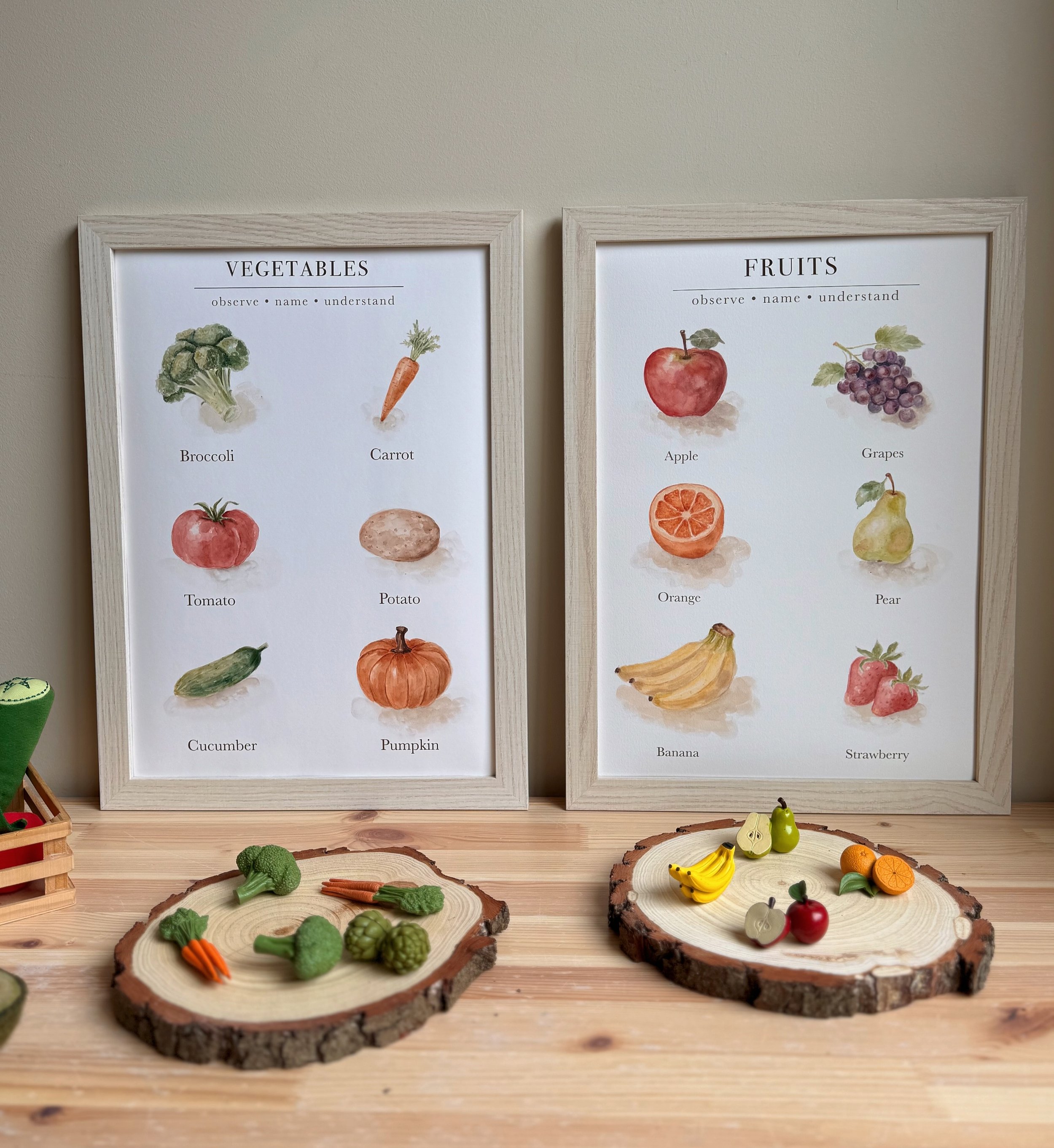 Educational display of toy fruits and vegetables, with two posters in the background labeled 'Vegetables' and 'Fruits' showing watercolor illustrations and names, with small wooden platforms holding additional toy produce on a wooden surface.