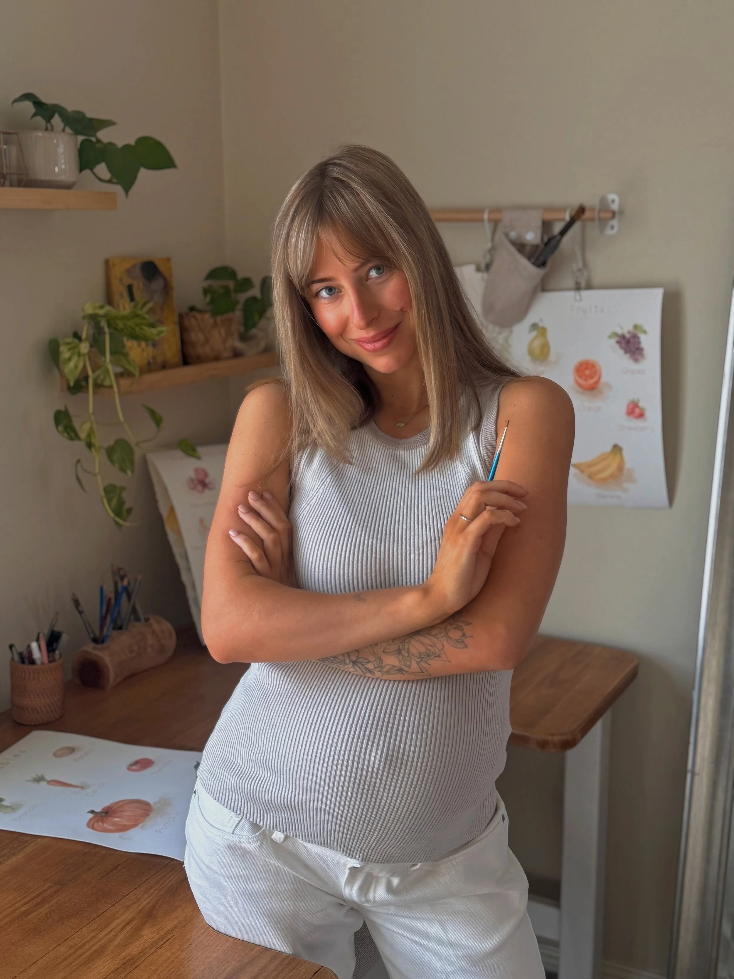 A woman with shoulder-length light brown hair, blue eyes, and a tattoo on her left forearm, smiling and looking at the camera, standing in a room with plants, artwork, and a wooden table.
