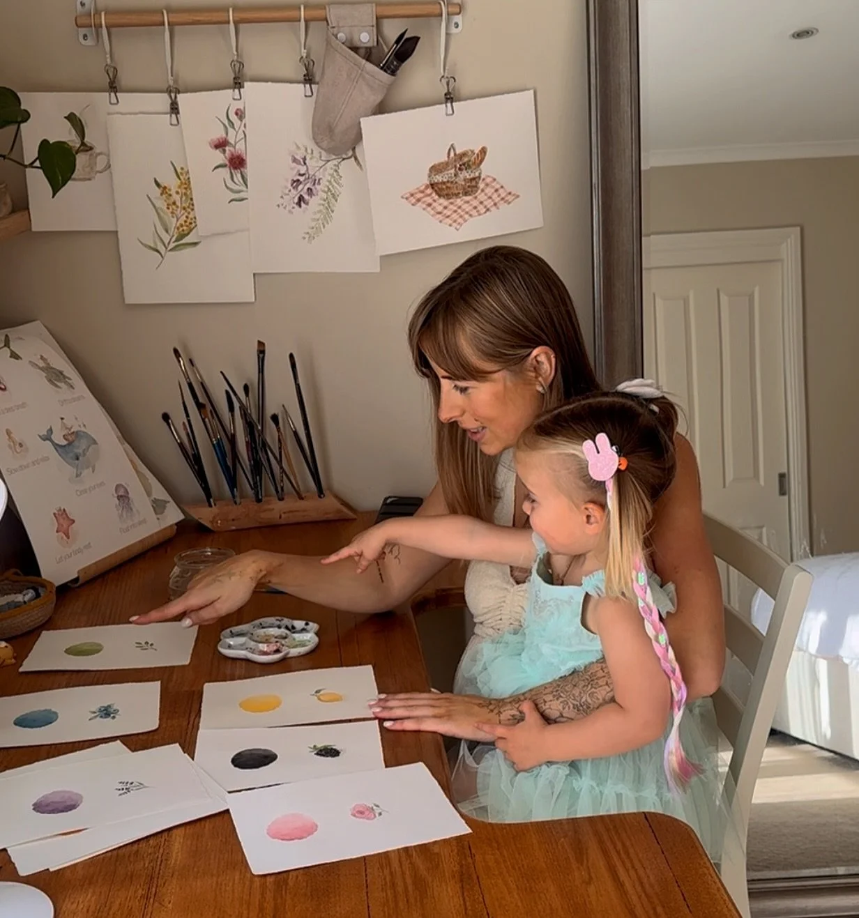 A woman and a young girl sit at a wooden table, engaging in arts and crafts with watercolor paintings of colorful circles and other designs. The girl points at her artwork, and the woman smiles helping her. The background includes watercolor paintings of flowers hanging on the wall and art supplies on the table.