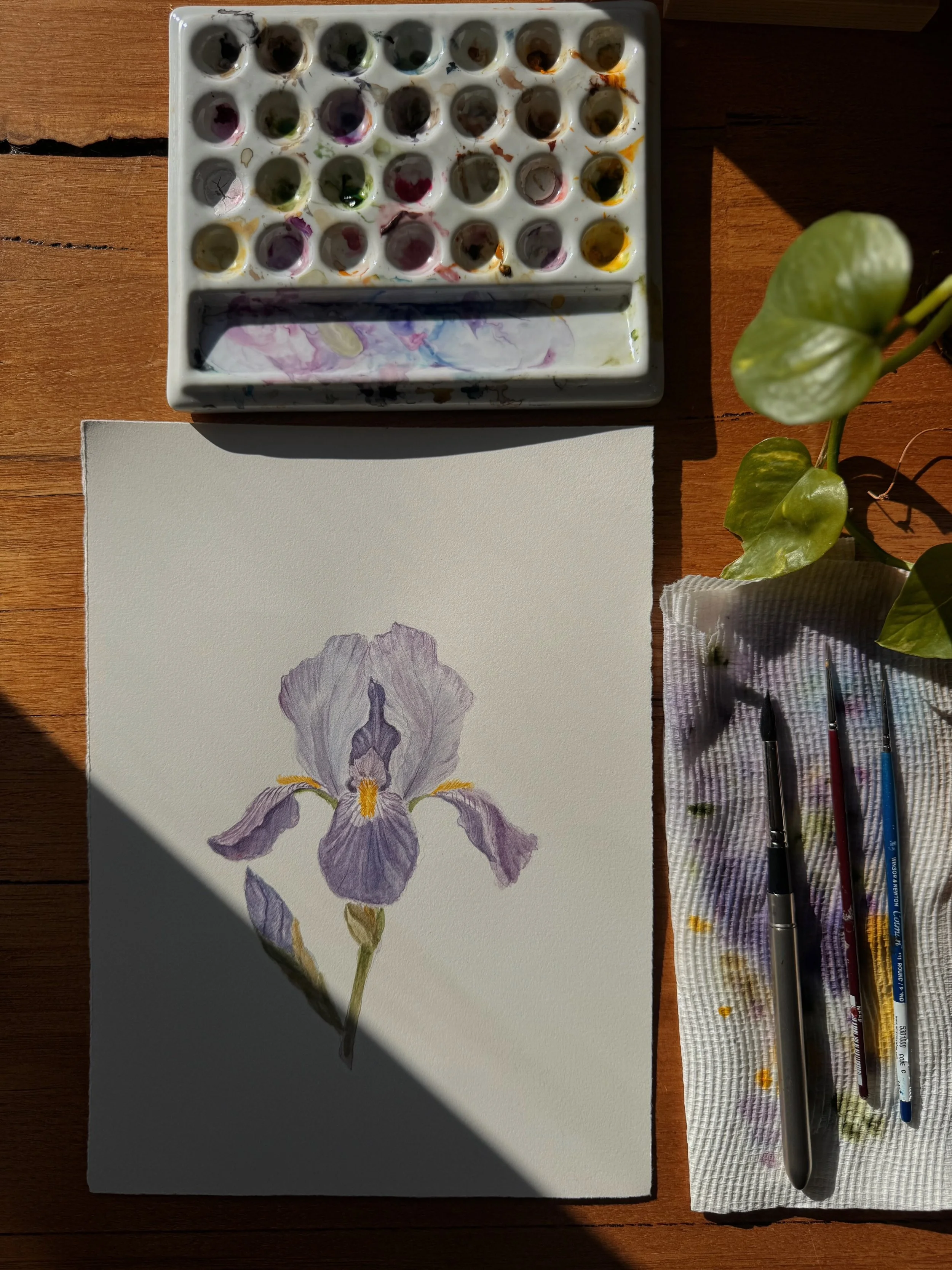A watercolor painting of a purple iris flower on a piece of watercolor paper, with watercolor paints, brushes, and a cloth with paint stains next to it on a wooden surface. sunlight and shadows are visible.