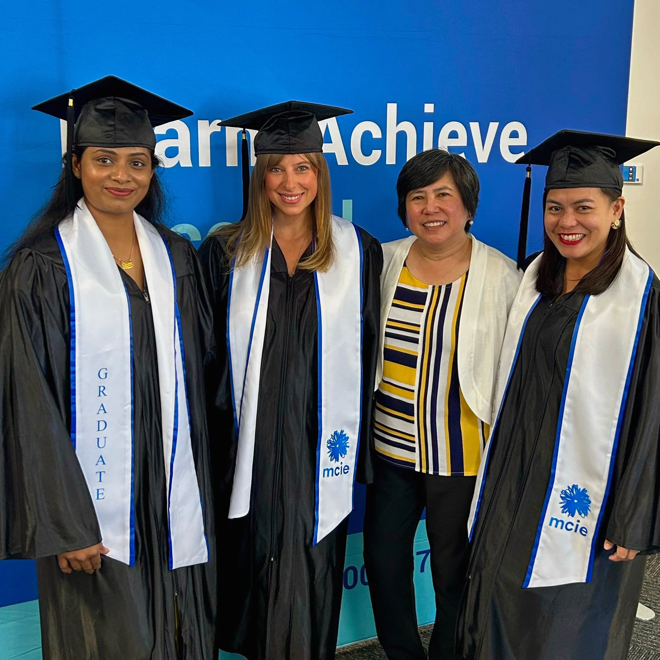 Group of four graduates in academic robes and caps standing in front of a blue sign with the words 'Learn Achieve' visible and smiling at the camera.