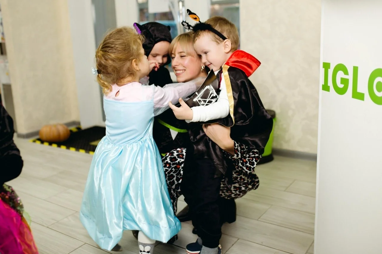Children dressed in Halloween costumes hugging a woman, celebrating at a party, with the word 'IGLO' partially visible in the background.
