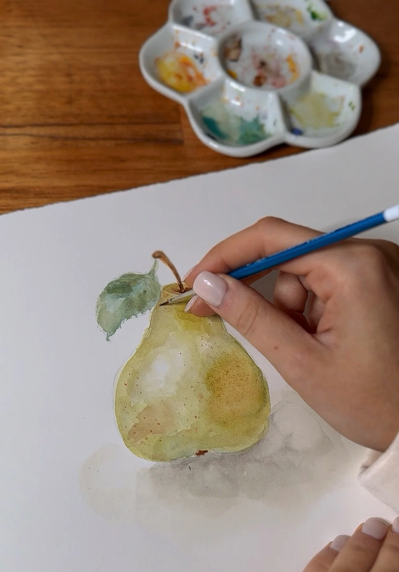 Watercolor painting of a yellow pear with green leaves on white paper, with a hand holding a paintbrush adding details.