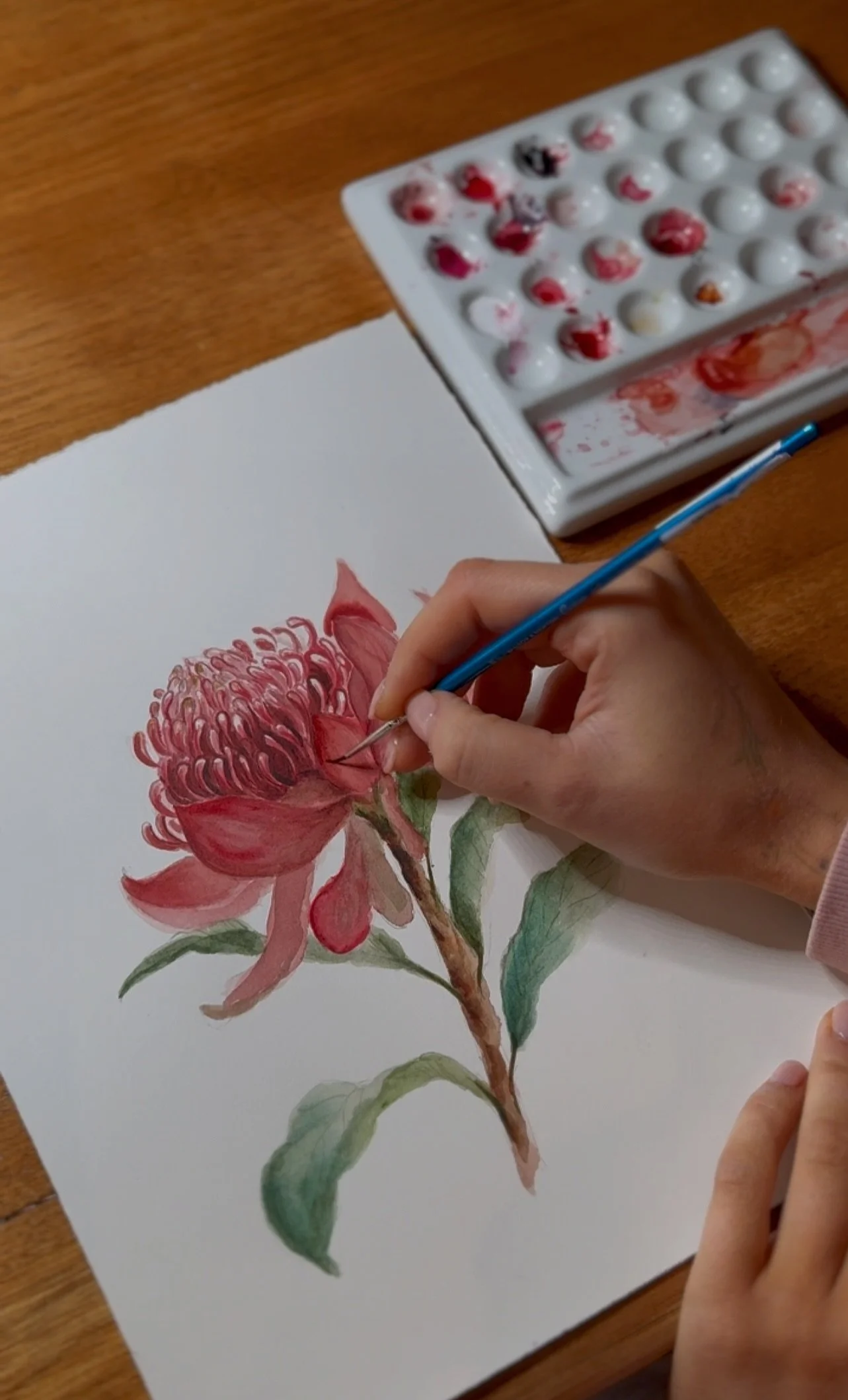 Person painting a detailed watercolor of a red flower with green leaves on white paper, with watercolor paints and brushes on a wooden table.