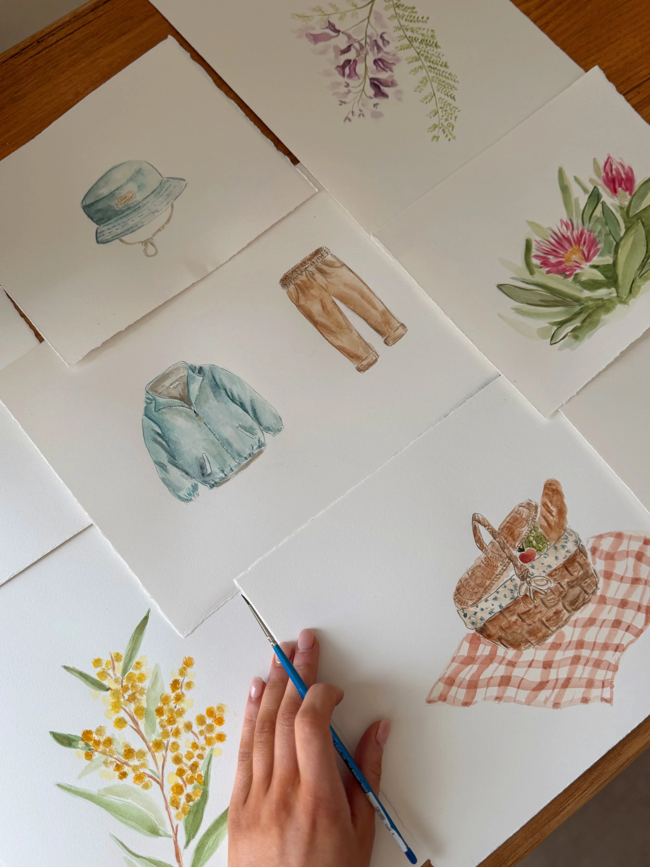 Watercolor illustrations of various objects including a blue hat, brown pants, a blue jacket, purple flowers, pink flowers, yellow flowers, and a woven basket with a red fruit and a gingham cloth.