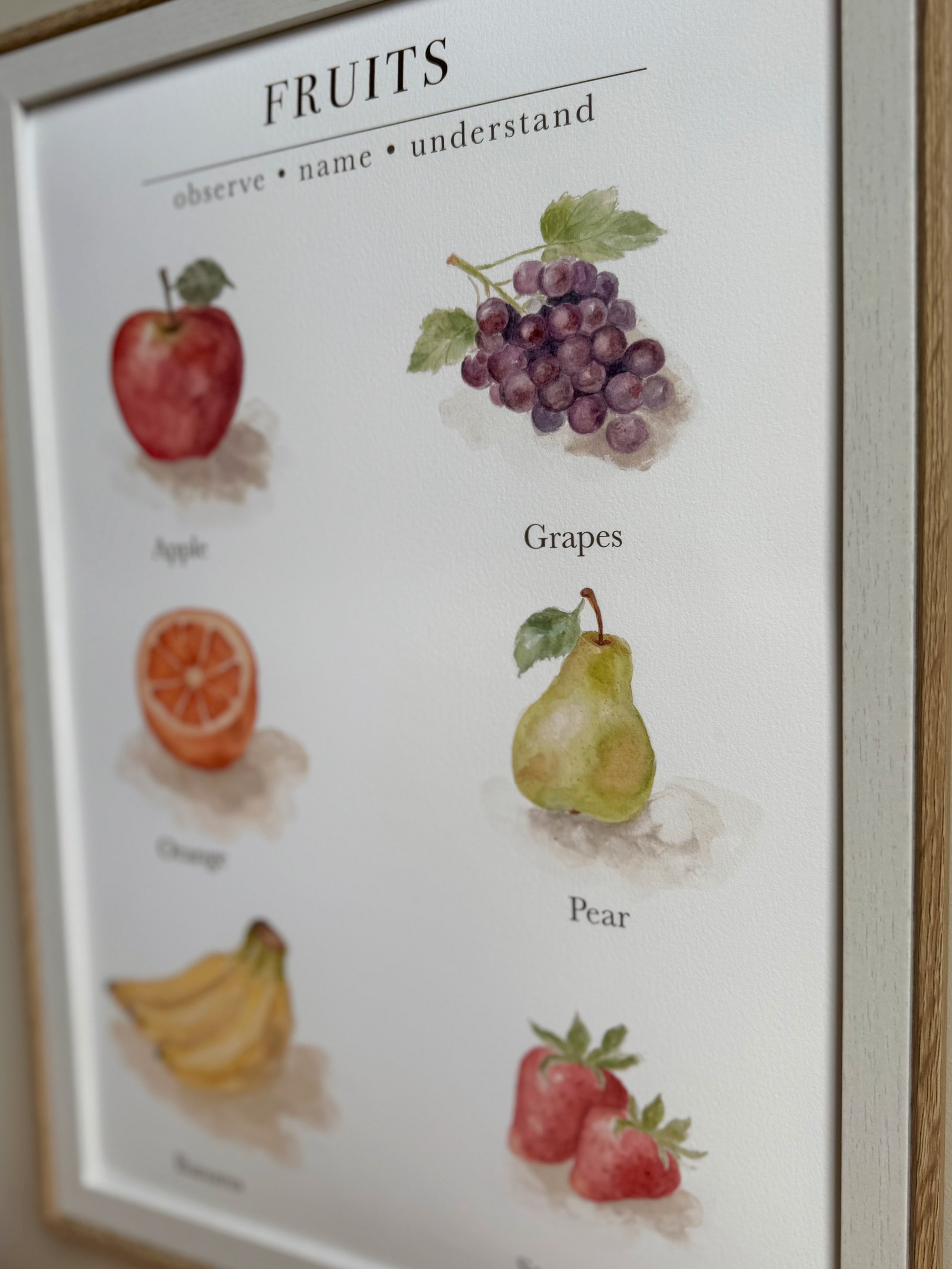 A framed poster displaying watercolor illustrations of various fruits, including grapes, apple, orange, pear, and strawberry, with their names labeled underneath.