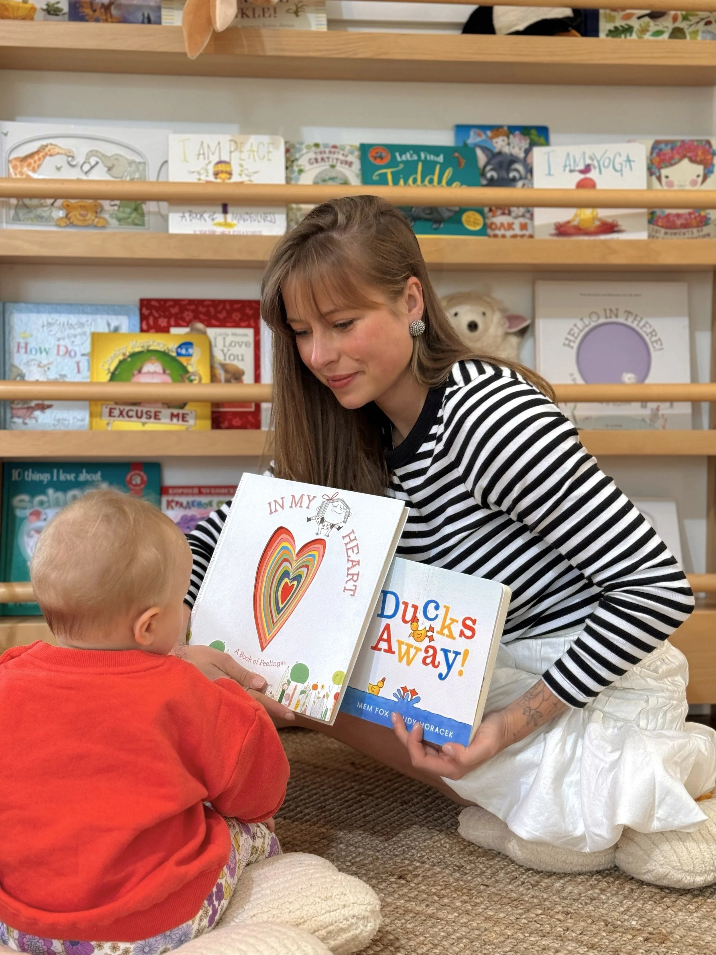 A woman with long brown hair, wearing a black and white striped shirt and white skirt, kneeling on a carpeted floor, reading a children's book to a young child with short blond hair wearing an orange shirt and patterned pants, in a bookstore with shelves filled with colorful children's books in the background.