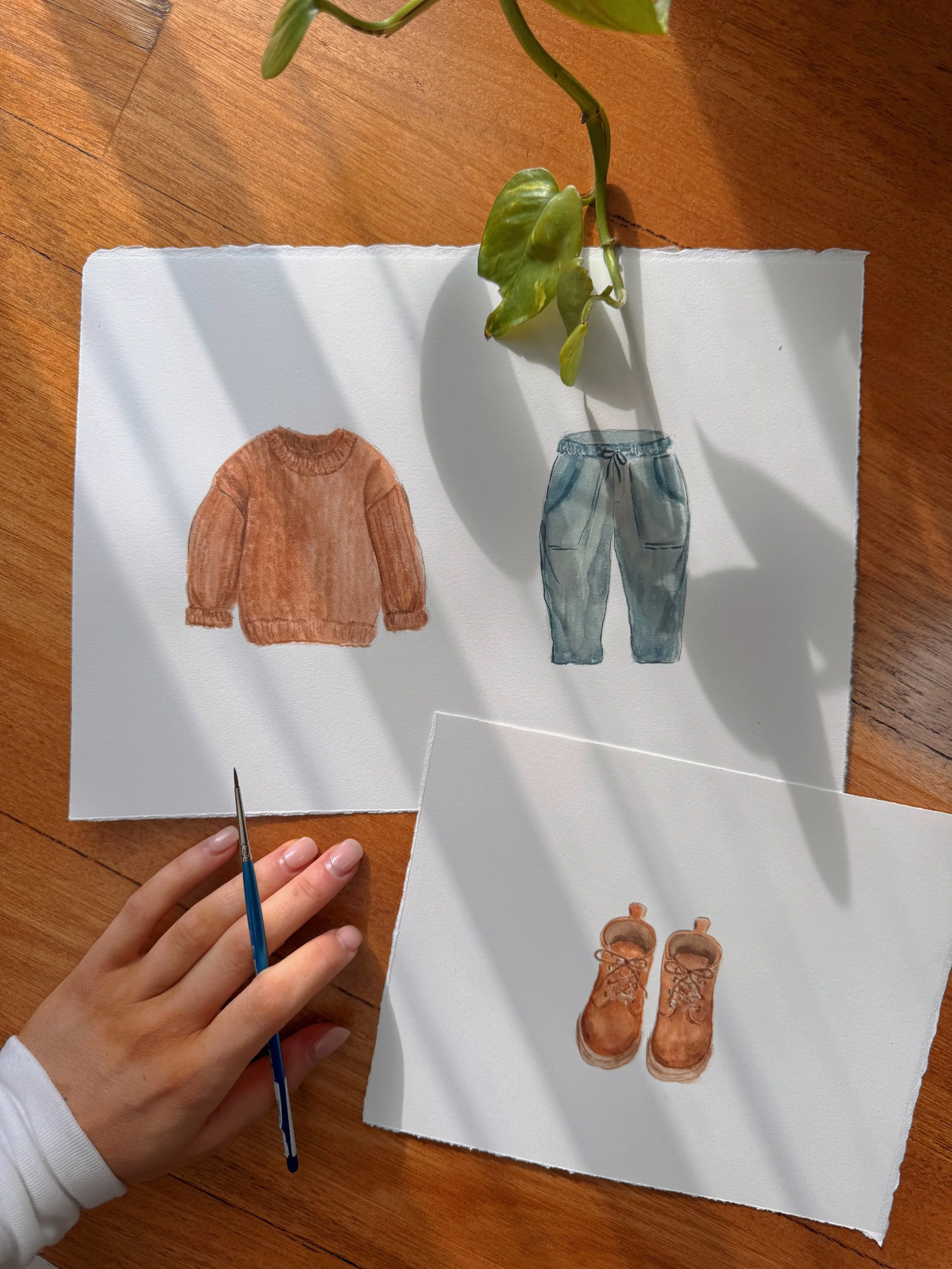 Watercolor illustrations of a brown sweater, gray jogger pants, and brown shoes on white paper, with a plant and sunlight shadows on a wooden table.