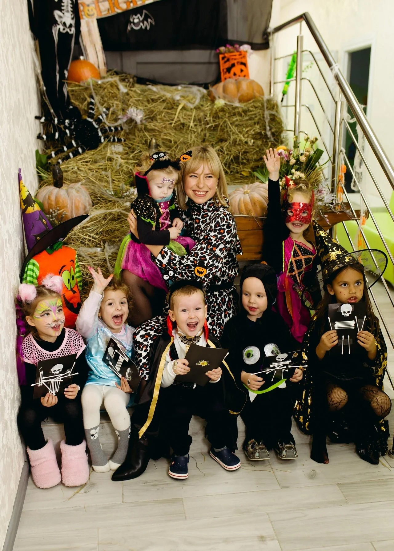 Group of children and a woman in Halloween costumes, sitting and standing on stairs, with Halloween decorations including pumpkins, spiders, and a spider web in the background.