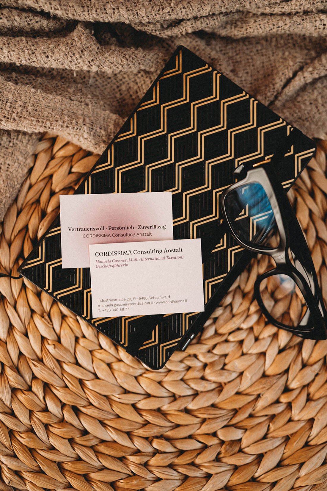 Arbeitsutensilien wie Manuela Gassners Brille, Visitenkarten, eine Notizbuch mit schwarzem und goldenem Muster, auf einem runden, geflochtenen Korbtisch, mit einem beige Stoff im Hintergrund.