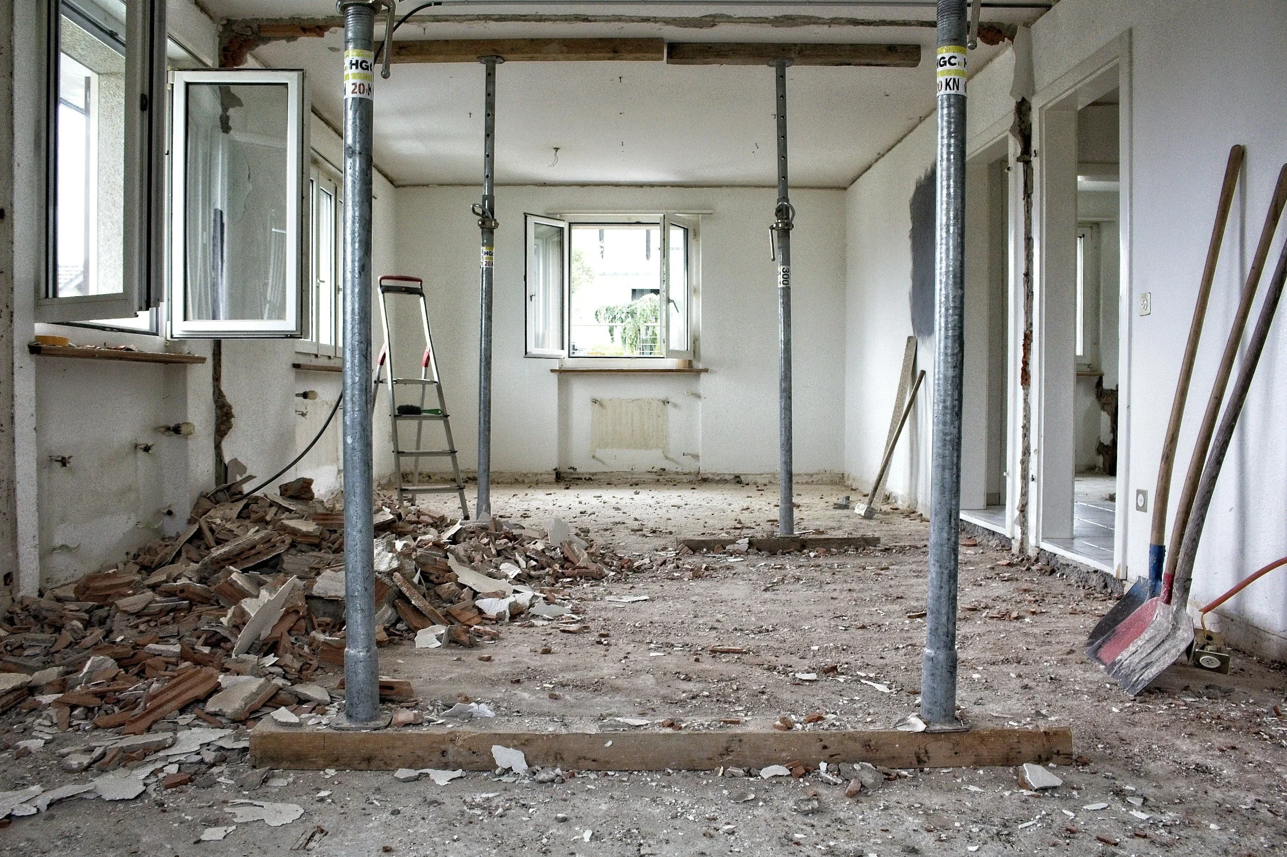 Interior of a room under renovation with scattered debris on the floor, exposed support beams, and open windows letting in natural light.