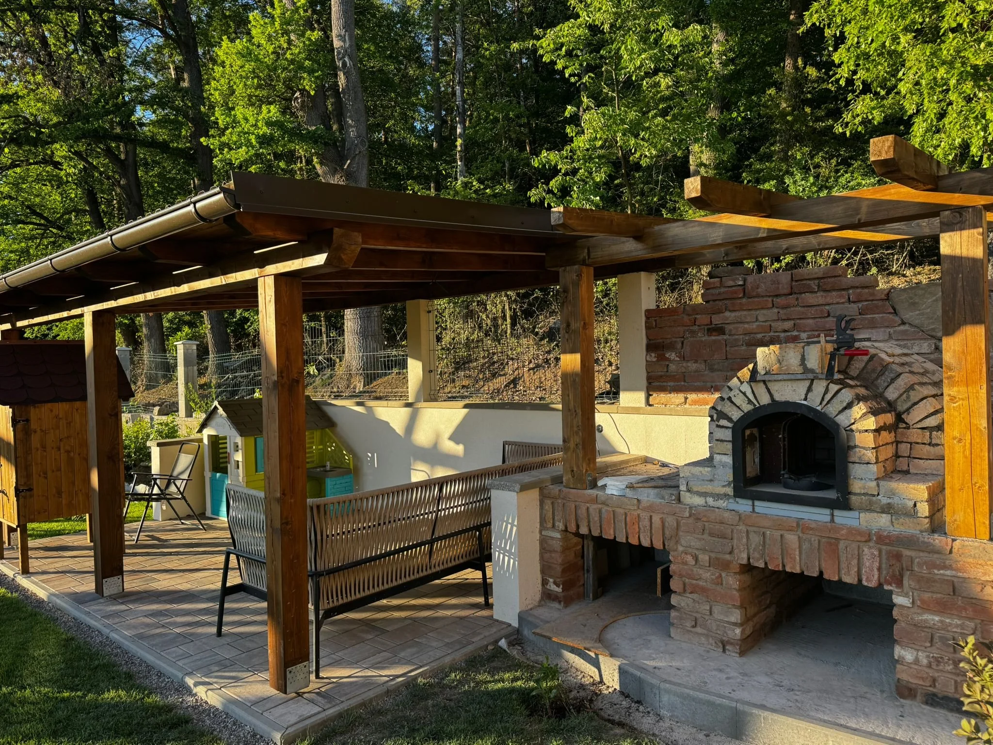 An outdoor patio area with a wooden pergola, a metal bench, a brick pizza oven, and surrounding trees and greenery.