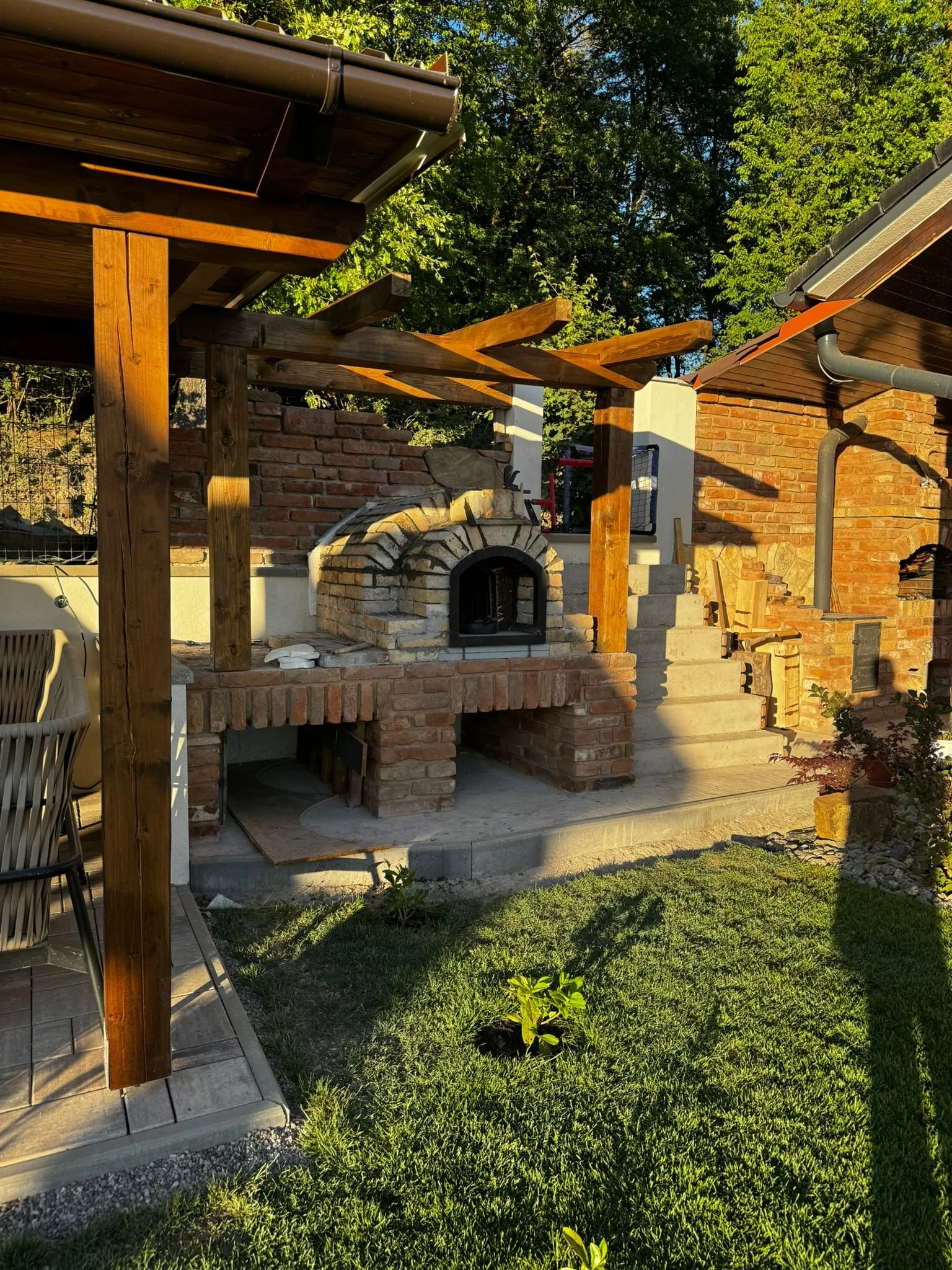 An outdoor patio with a brick pizza oven, a wooden pergola, a brick wall, and green grass and trees in the background.