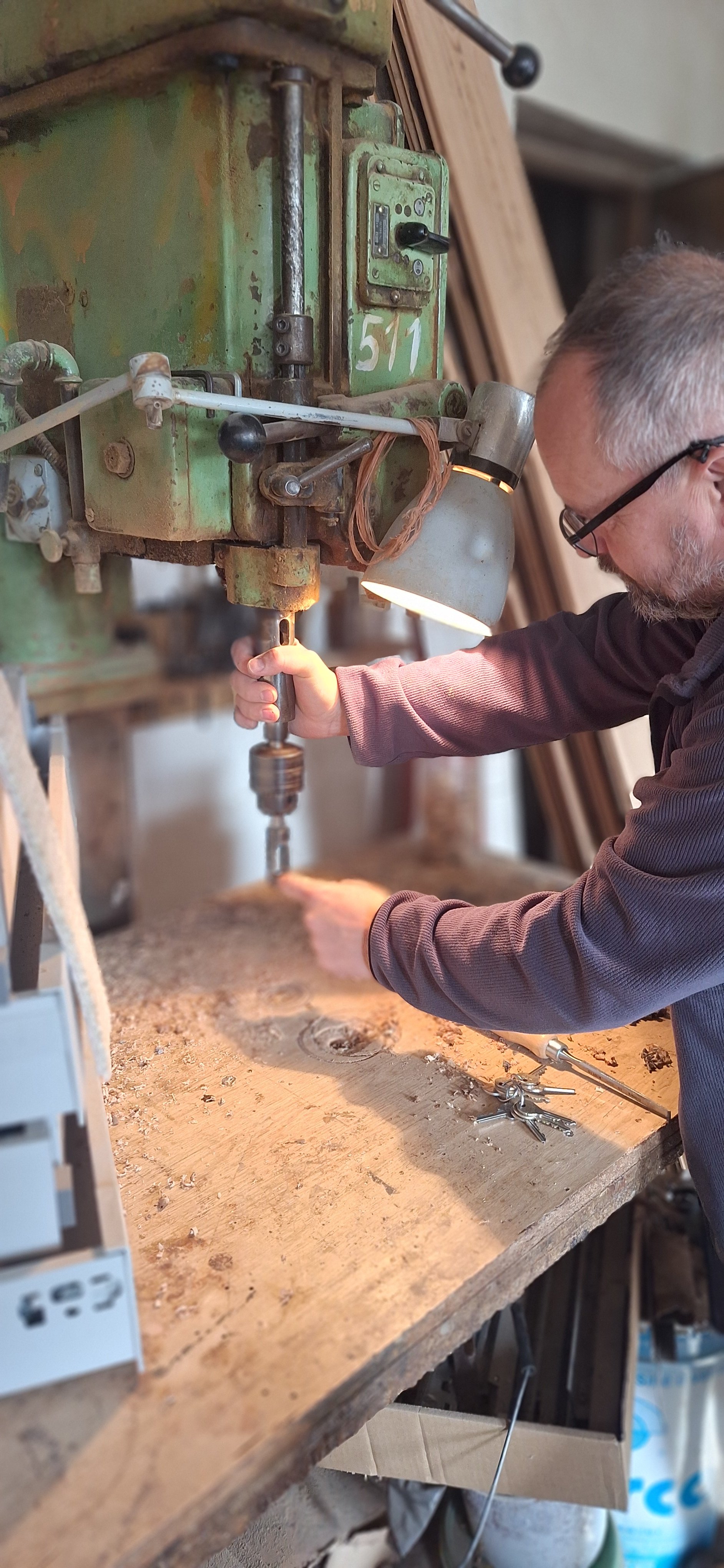 A man works on a green industrial drill press, drilling into a piece of wood on a workbench in a workshop.