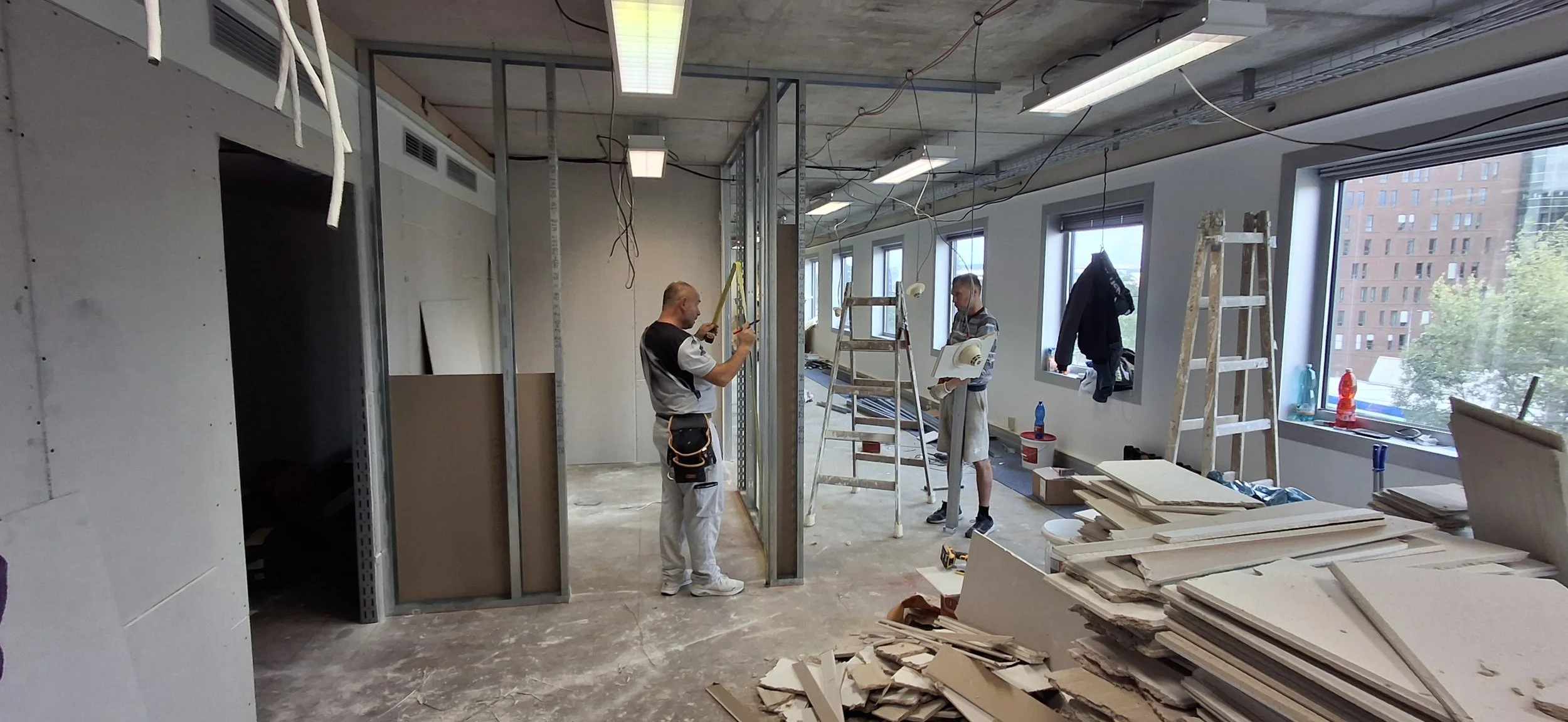 Construction workers installing metal framing for interior walls in an office building under renovation, with tools and materials around.