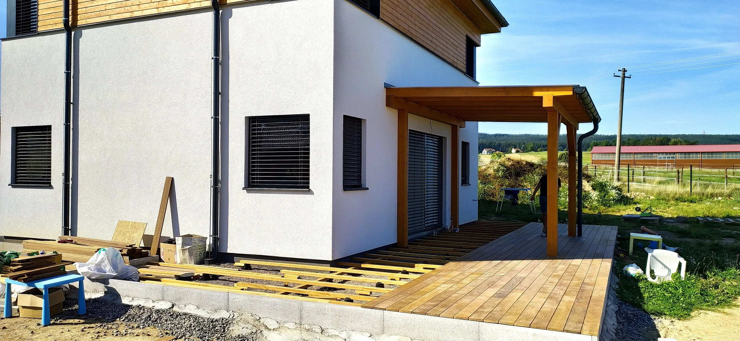 Newly constructed house with a wooden patio and outdoor area under construction, with tools and materials scattered around, in a rural setting with open fields and a barn in the background.