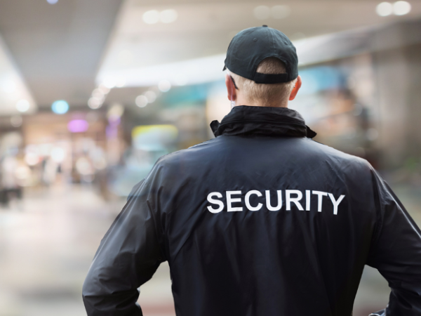 A security guard with short blond hair, wearing a black jacket with 'SECURITY' printed on the back and a black cap, standing in a busy indoor shopping mall.